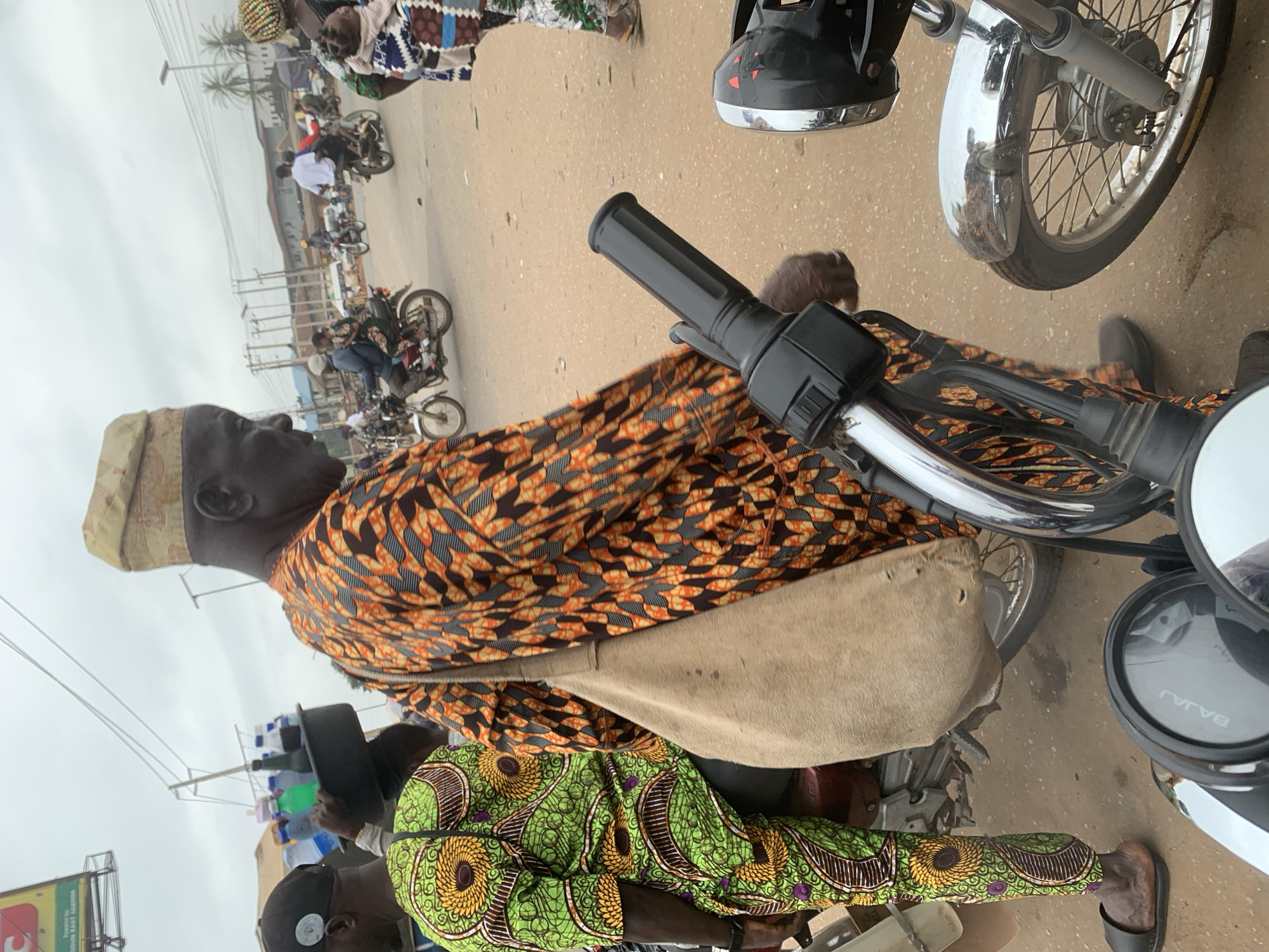 The image depicts a street scene in Shagamu, Nigeria. In the foreground, a man wearing a traditional orange and black patterned dashiki and a matching cap is seen from the side. He has a large, light brown woven bag slung over his shoulder. To his left, another person, partially visible, wears a green and yellow patterned dashiki and pants, also with a black cap. The man in the orange dashiki is positioned next to a motorcycle, with the handlebars and front wheel of another motorcycle visible in the immediate foreground. The background reveals a dusty road with several motorcycles and people. In the mid-ground, motorcycles are in motion, with riders and passengers. Further back, buildings and power lines suggest an urban environment. The sky is overcast. The scene appears to be a busy marketplace or street intersection.