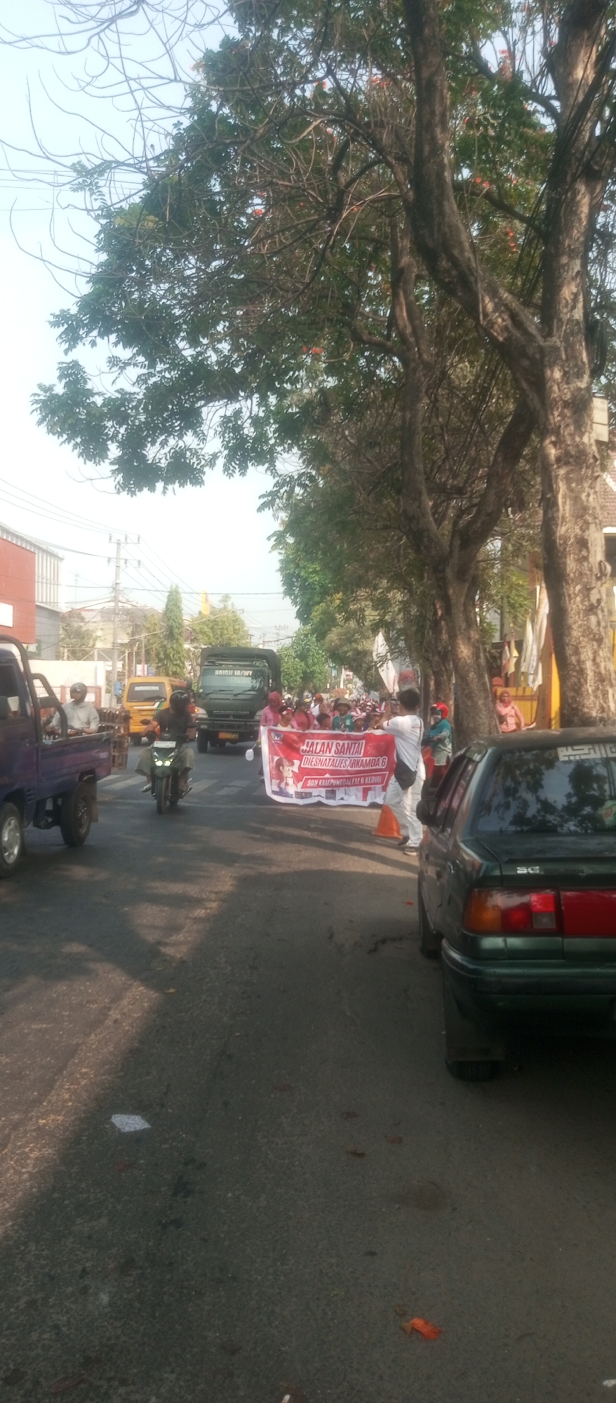 In the city of Kediri, Indonesia, on a paved road lined with trees, a procession is underway. A large red banner with white text reads "JALAN SANTAI DIESNATALIES ARKAMDA 6 SON KAMPINGDALEM 6 KEDIRI". Several people are visible holding the banner and walking, some in the distance. A green truck is in the background, and a yellow pickup truck is to its left. A motorcycle is in motion on the road, with a rider. To the right, the rear of a dark green car is visible. The sky is clear and the sun is casting shadows on the road. The environment suggests a public event or parade taking place on a street.