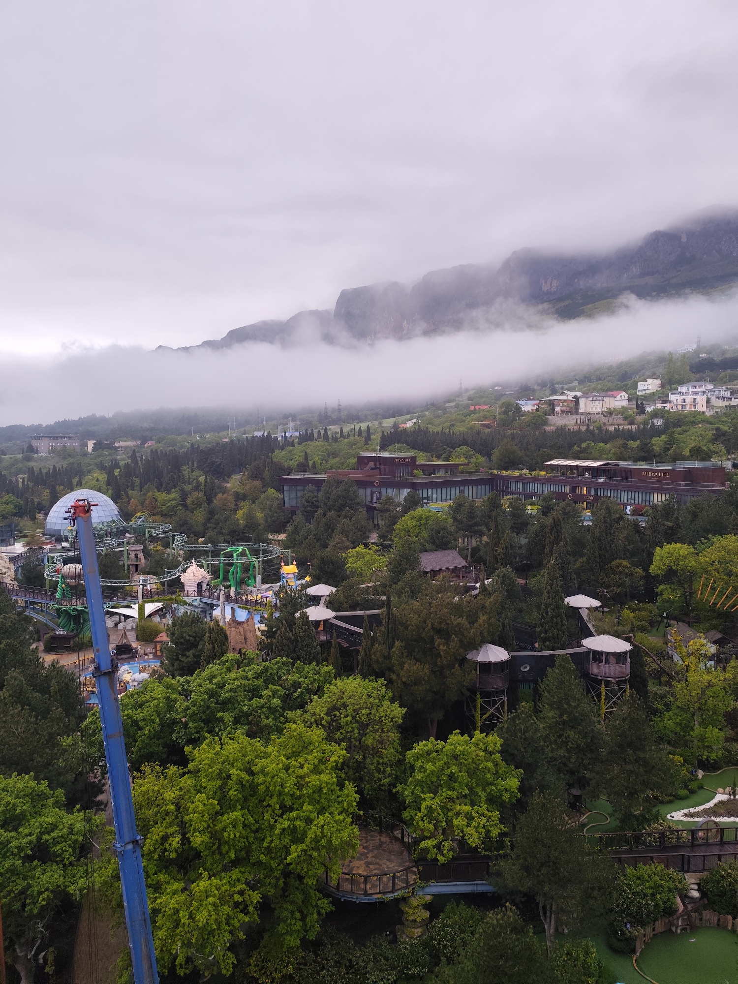 The image depicts a view in Симеизский поселковый совет, Ukraine, characterized by a blend of natural and developed elements under an overcast sky.
In the middle ground, a large amusement park sprawls amidst lush greenery, with visible rides, including a roller coaster, and various structures painted in bright colors. A blue support structure for a ride stands prominently in the foreground. The park merges into dense tree cover which gives way to residential or commercial buildings, including a long, multi-story hotel complex with "Mriya Life" inscribed on its facade.
The backdrop features a rugged mountain range partially obscured by low-hanging clouds and fog. The heavy cloud cover diffuses the light, lending a soft, muted tone to the overall scene. The atmosphere appears damp and cool.
