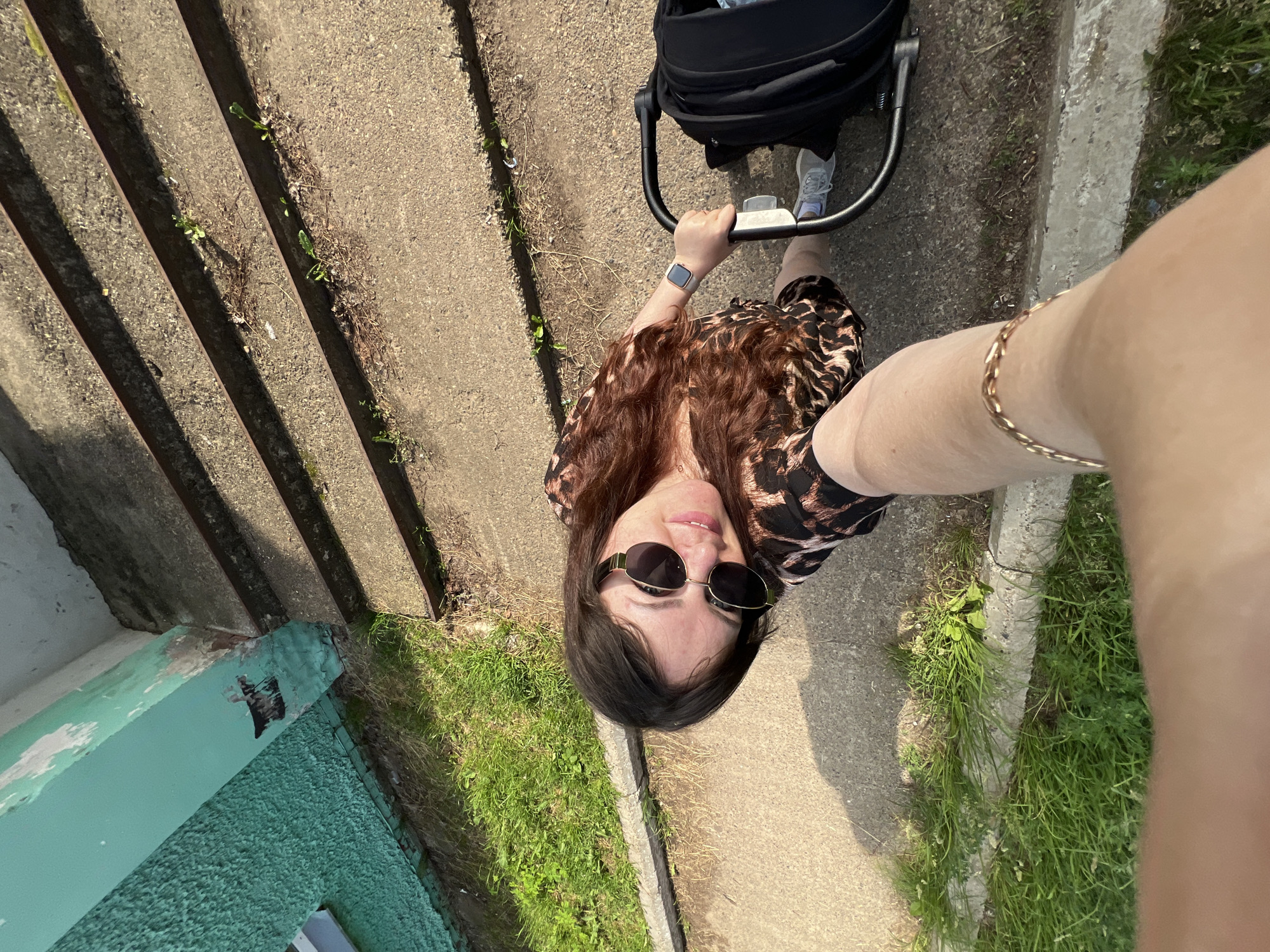 A woman is taking a selfie while pushing a stroller on a sunny day. She is wearing a leopard-print dress, sunglasses, and a gold bracelet. The stroller is black. The scene appears to be outdoors, possibly in front of a building with teal-colored paint. There are concrete stairs and patches of grass visible. The location is identified as Noviy, Russia.