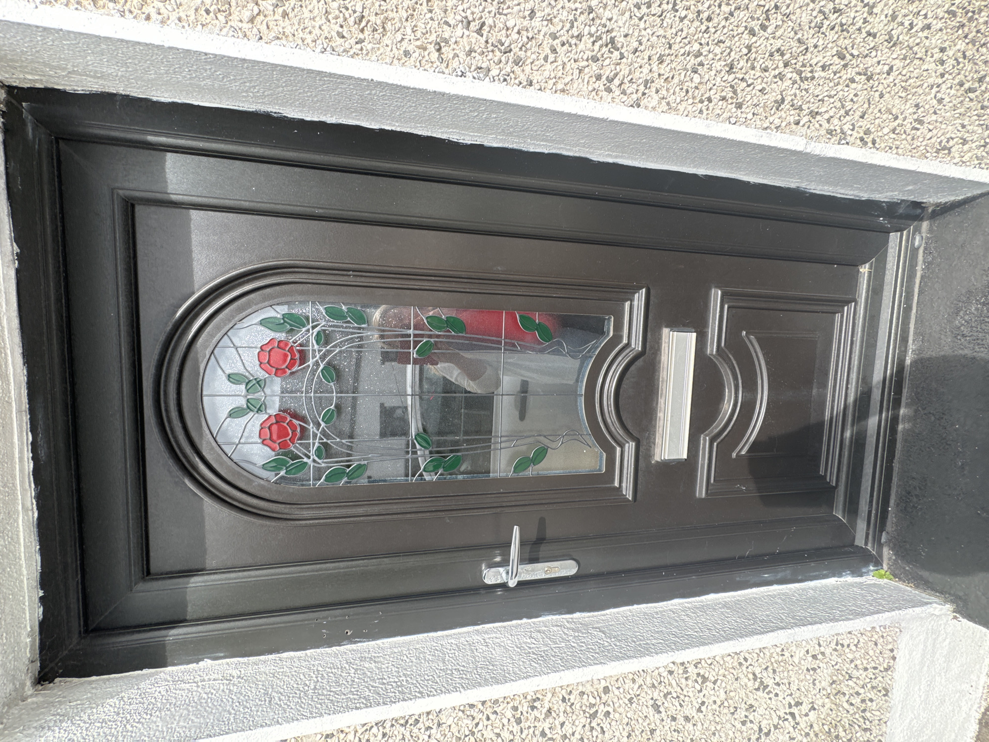 The image shows a dark grey front door with stained glass featuring red roses and green leaves. A silver door handle and keyhole are visible on the left side of the door, and a silver mail slot is centered below the stained glass. The door is set within a textured white frame and is adjacent to a rough, speckled wall. A reflection in the stained glass shows a person wearing a red top. The scene appears to be an exterior view of a residential property. The specific location in Waterford, Ireland, is not discernible from the image alone.