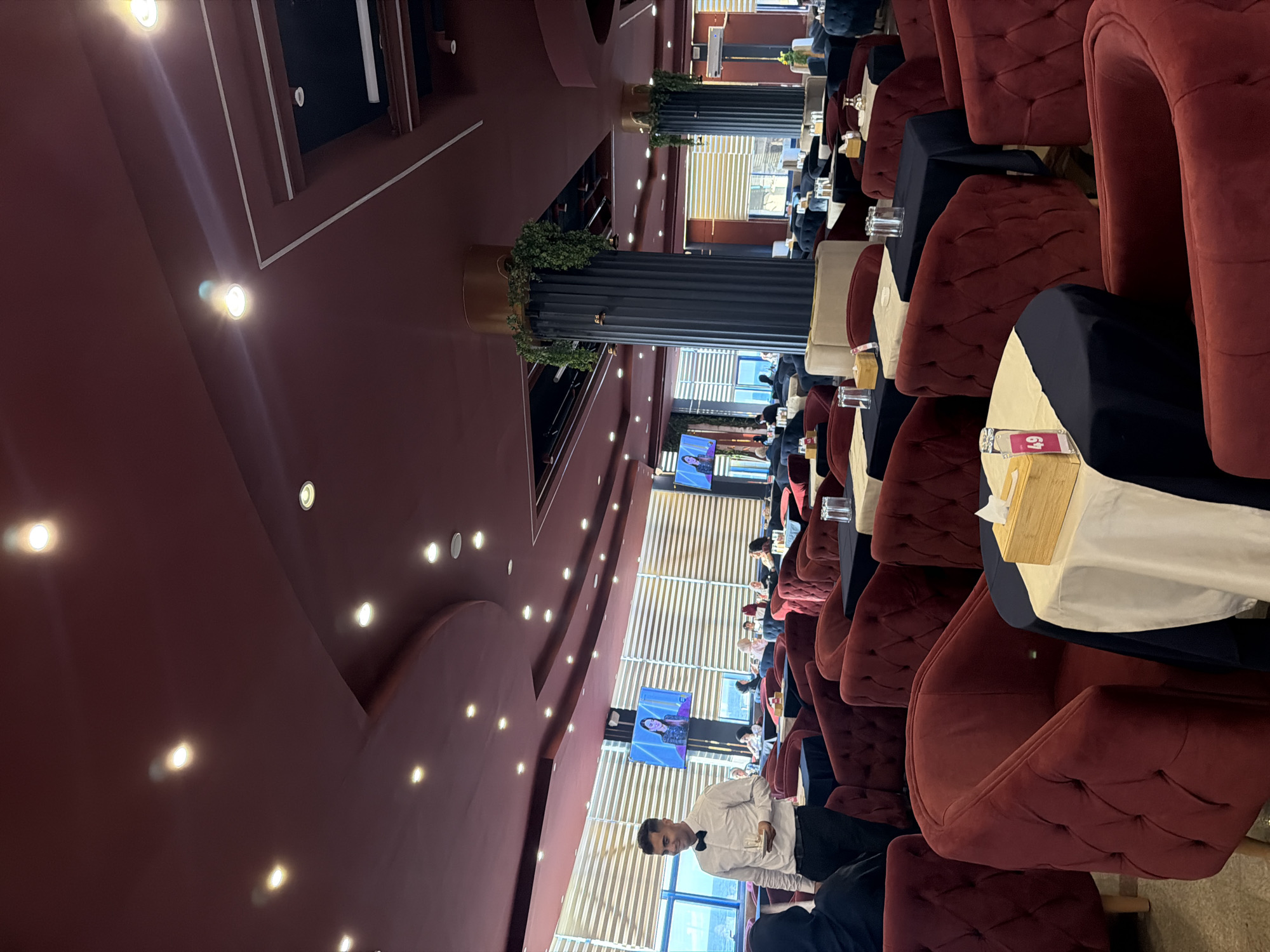The image shows the interior of a spacious cafe or lounge located in the city of Unknown, Unknown, featuring large windows overlooking a body of water and a distant landscape. The ceiling is a dark burgundy color, illuminated by numerous recessed spotlights, and incorporates curved architectural elements. Two prominent, dark gray fluted columns, topped with gold-colored bands and adorned with artificial green foliage, are situated within the space. Seating arrangements include numerous deep red, upholstered armchairs with tufted backs and some lighter beige upholstered low partition walls. Tables are covered with dark blue tablecloths and lighter cream-colored runners. In the foreground, a male waiter, dressed in a white shirt, black bow tie, and black trousers, stands holding a glass on a saucer, smiling towards the left. Several patrons are visible seated at tables in the background. Two large flat-screen televisions are mounted on columns, each displaying a woman. One table in the foreground has a wooden tissue box and a small pink sign with the number '49'. An air conditioning unit is mounted high on a wall in the background.