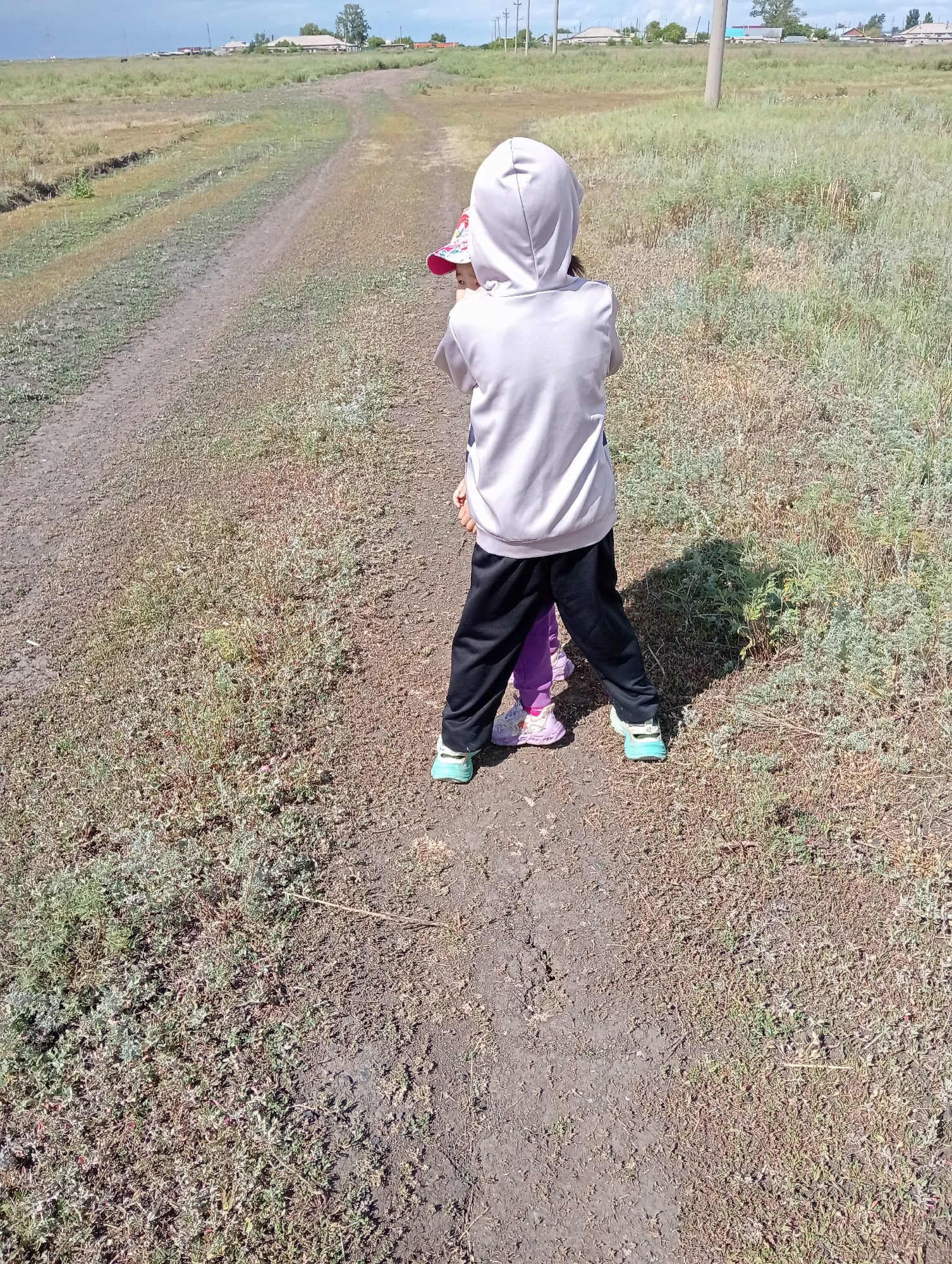 Two children stand on a dirt road in a rural area. The child in the foreground, wearing a light-colored hoodie, black pants, and light blue sneakers, is facing away from the camera. A second child, partially visible beneath the first, wears purple pants and pink and white sneakers. They are in a field with dry grass and sparse vegetation, with a dirt path winding into the distance. In the background, several houses and utility poles are visible under a cloudy sky.