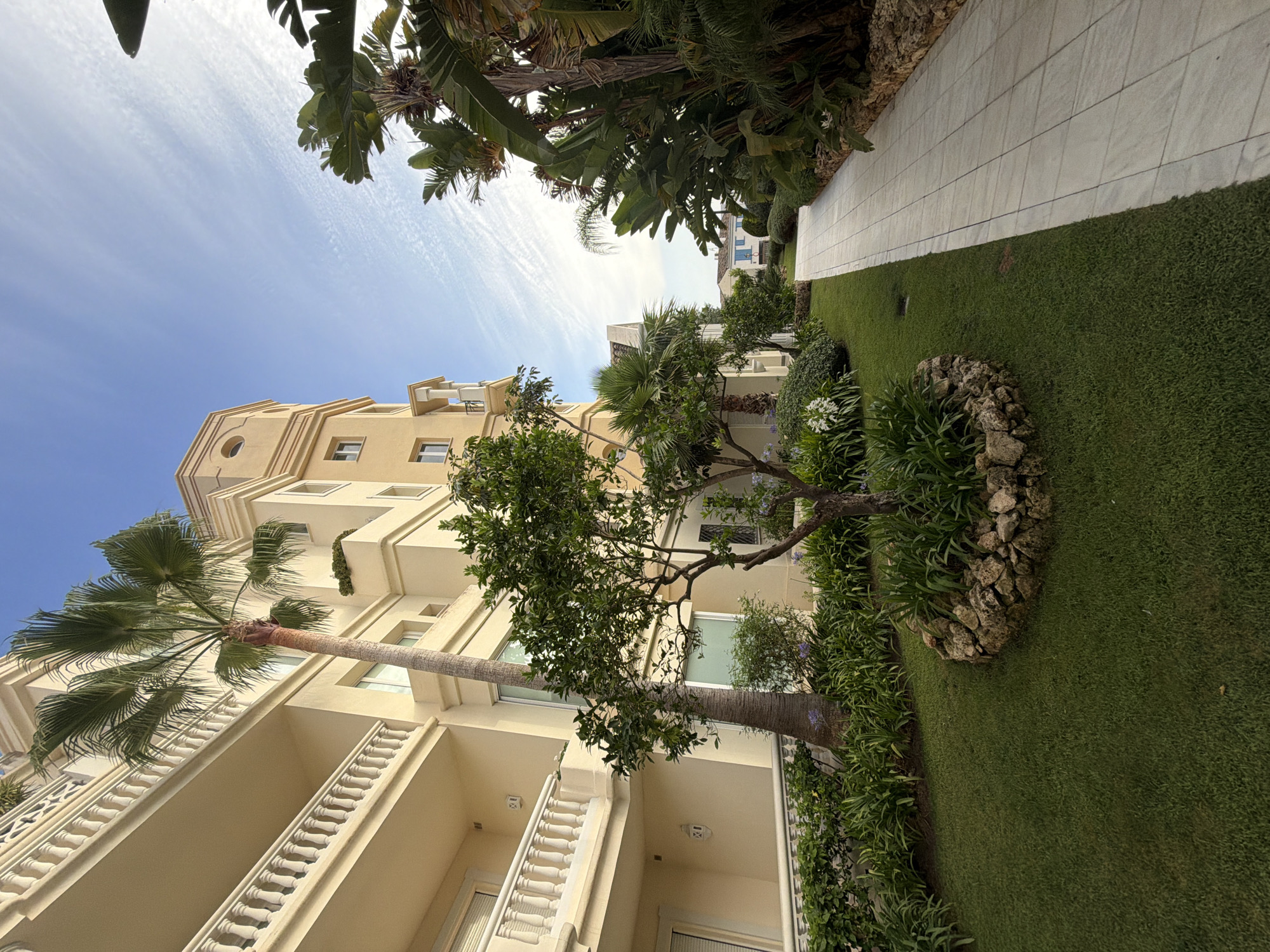 The image depicts a scene in the city of Resinera-voladilla, Spain. A modern apartment building with cream-colored facade and balconies with white railings dominates the left side of the frame. In front of the building, there is a well-maintained garden with lush green grass, a small tree surrounded by rocks, and various plants and palm trees. A paved walkway runs along the right side of the garden, bordered by more tropical vegetation and rocks. The sky is blue with wispy white clouds. The overall atmosphere is serene and luxurious, suggestive of a resort or upscale residential area. There are no people or animals visible in the image.
