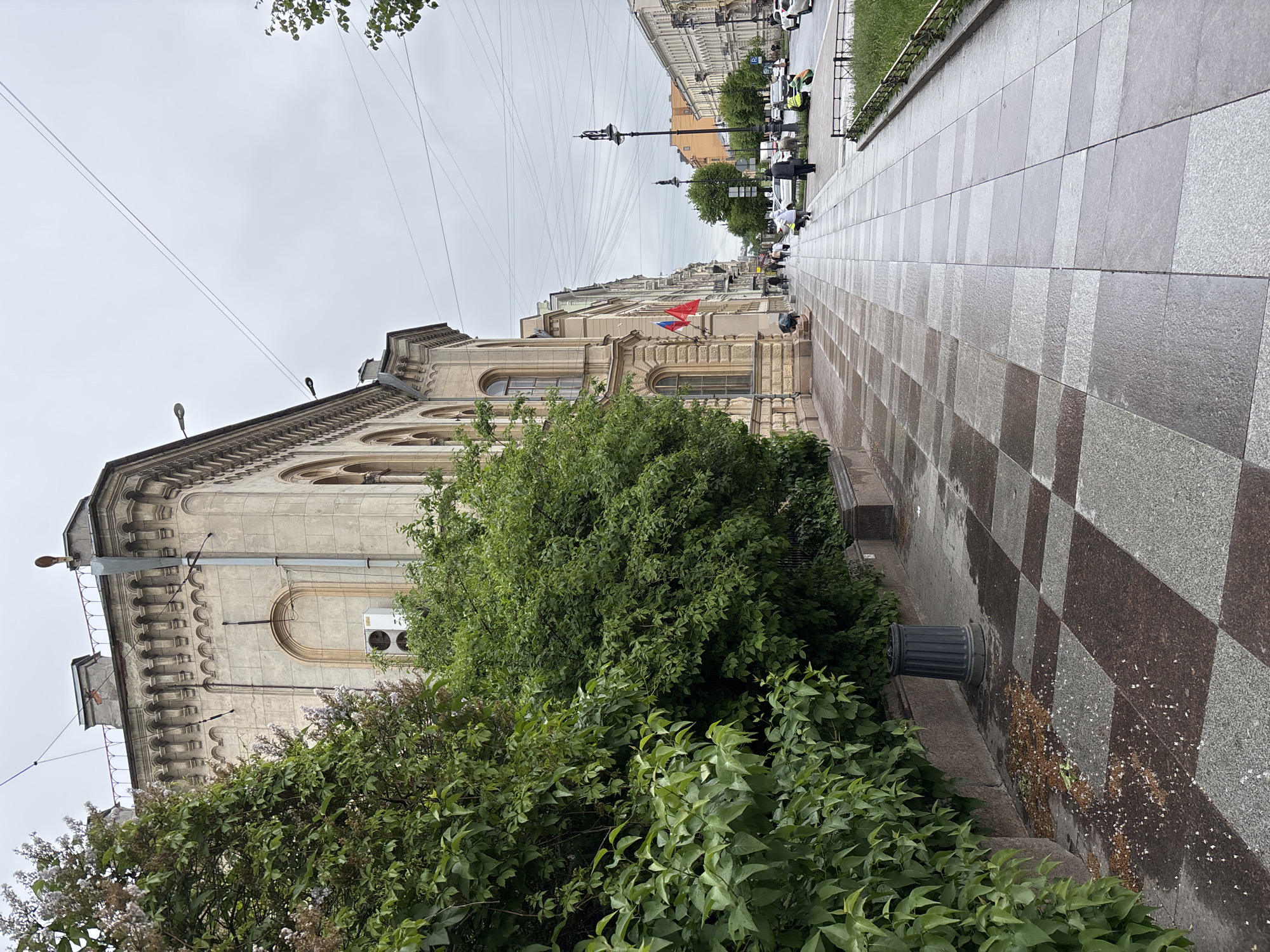 The image captures a street scene in Saint Petersburg, Russia, characterized by overcast skies and a wet, reflective sidewalk. The architecture is notable, with a large, historical building on the left side of the frame. The building exhibits an ornate facade with arched windows, a light-colored exterior, and what appears to be a Russian flag displayed. In the foreground, there is dense green foliage obscuring part of the building's base. The sidewalk is paved with large, gray and brown checkered tiles. In the distance, the street extends, lined with buildings, trees, street lamps, and some vehicles. People are walking along the sidewalk, with some appearing to be participating in some sort of street cleaning or maintenance. Overhead, there are numerous electrical wires crisscrossing the sky.