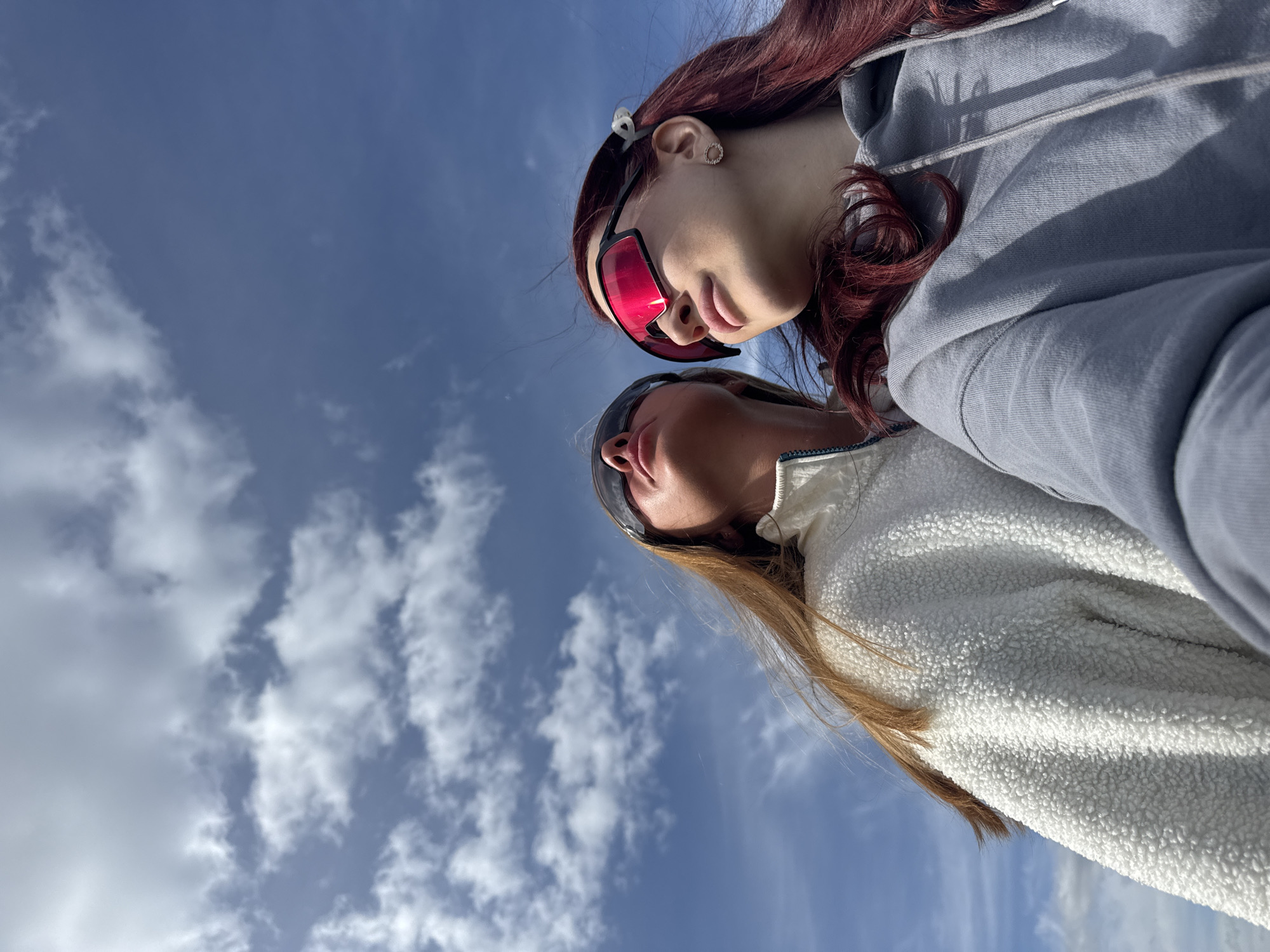 Two women are standing close together, likely taking a selfie, with a low-angle perspective capturing their faces and the sky behind them. The woman in the foreground has long, light brown hair and is wearing a white fuzzy jacket and sunglasses. The woman behind her has long, red hair and is wearing a gray hoodie and red sunglasses. The sky is blue with scattered white clouds. The location is Bol'shoye Kozino, Russia.