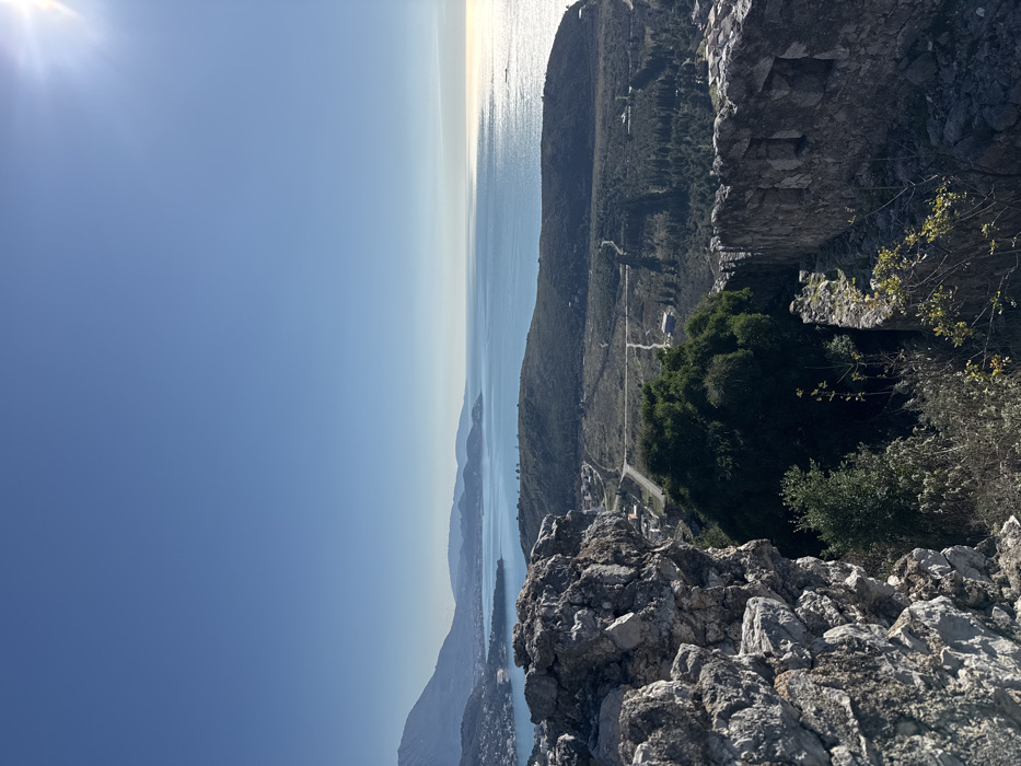 The image displays a panoramic, high-angle view overlooking a coastal landscape from within ancient stone ruins in Zankovići, Montenegro. In the immediate foreground, to the left, are large, weathered grey stone walls and rock formations indicative of an old fortification, with smaller green shrubs and sparse vegetation growing amongst the crevices. To the right, further stone ruins, possibly part of a wall with an opening, are visible, also overgrown with sparse green plants.
The midground reveals a rolling terrain descending towards the coast, characterized by dry, light-colored vegetation interspersed with darker green trees and patches of dense foliage. A winding paved road traverses this terrain, connecting scattered small buildings and structures.
The background features the deep blue expanse of the Adriatic Sea, reflecting sunlight brightly on its surface towards the right, creating a distinct sun glare. Along the left coastline, a bay is visible, fronting a coastal city (likely Bar) with numerous white and light-colored buildings clustered against the base of large, verdant mountains that rise abruptly from the sea. Distant landforms and hazy mountain ranges extend along the horizon. The sky above is clear and bright blue, with the sun prominently positioned in the upper right quadrant. No individuals, animals, or active interactions are present in the scene. The environment is outdoors, under clear skies, on what appears to be a sunny day.