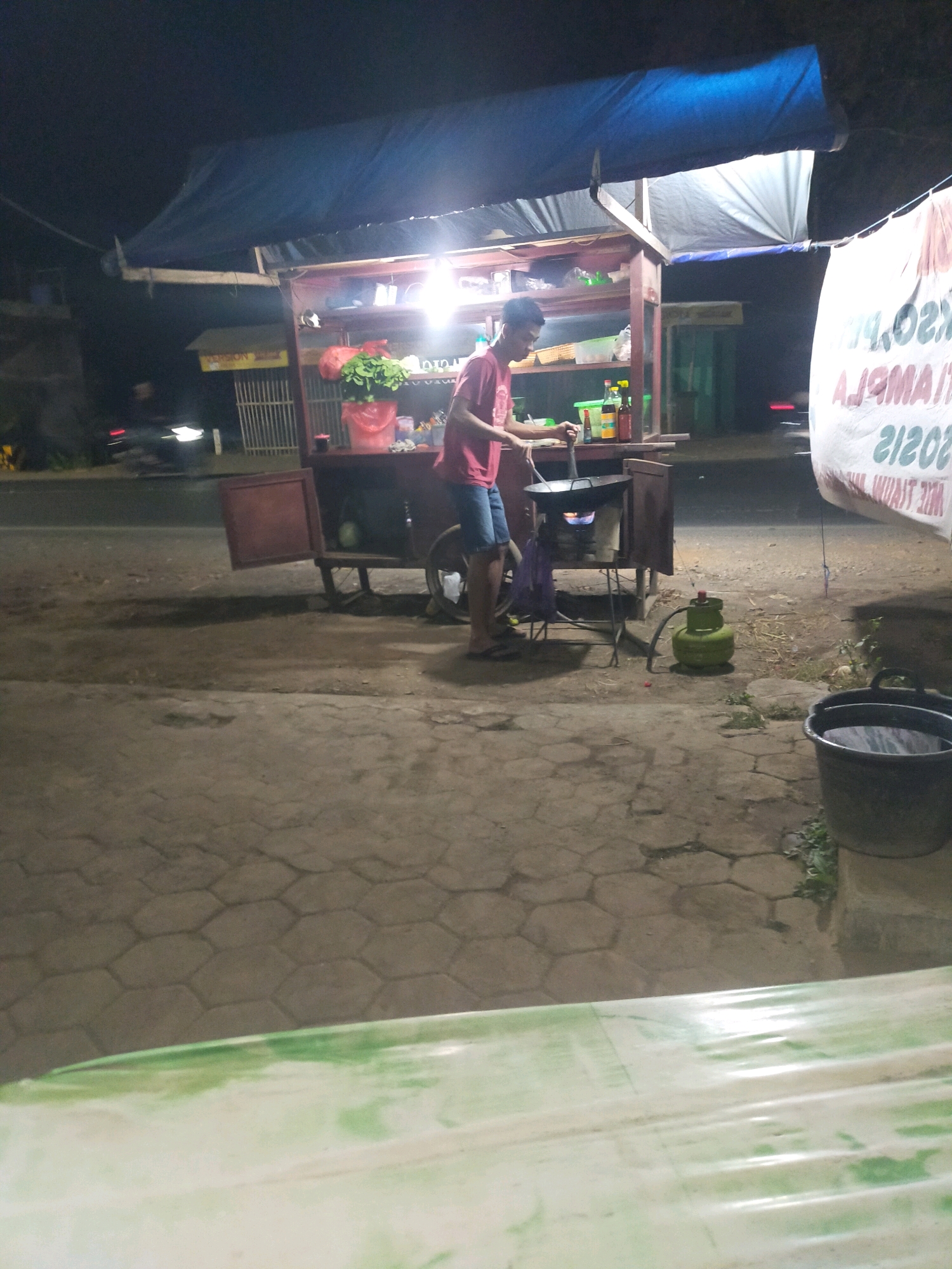 A man, wearing a red t-shirt and blue shorts, stands in front of a food cart at night. The cart has a blue tarp overhead and is lit by overhead lights. There are shelves within the cart displaying food items and condiments. A wok is positioned over a gas burner on a stand in front of the cart, and the man appears to be cooking. A green propane tank is connected to the burner. A large white banner with green writing is hanging on the right side of the image. In the background, a road is visible with a car driving past. The ground is paved with hexagonal tiles. The scene appears to be in Gedong Wetan, Indonesia.