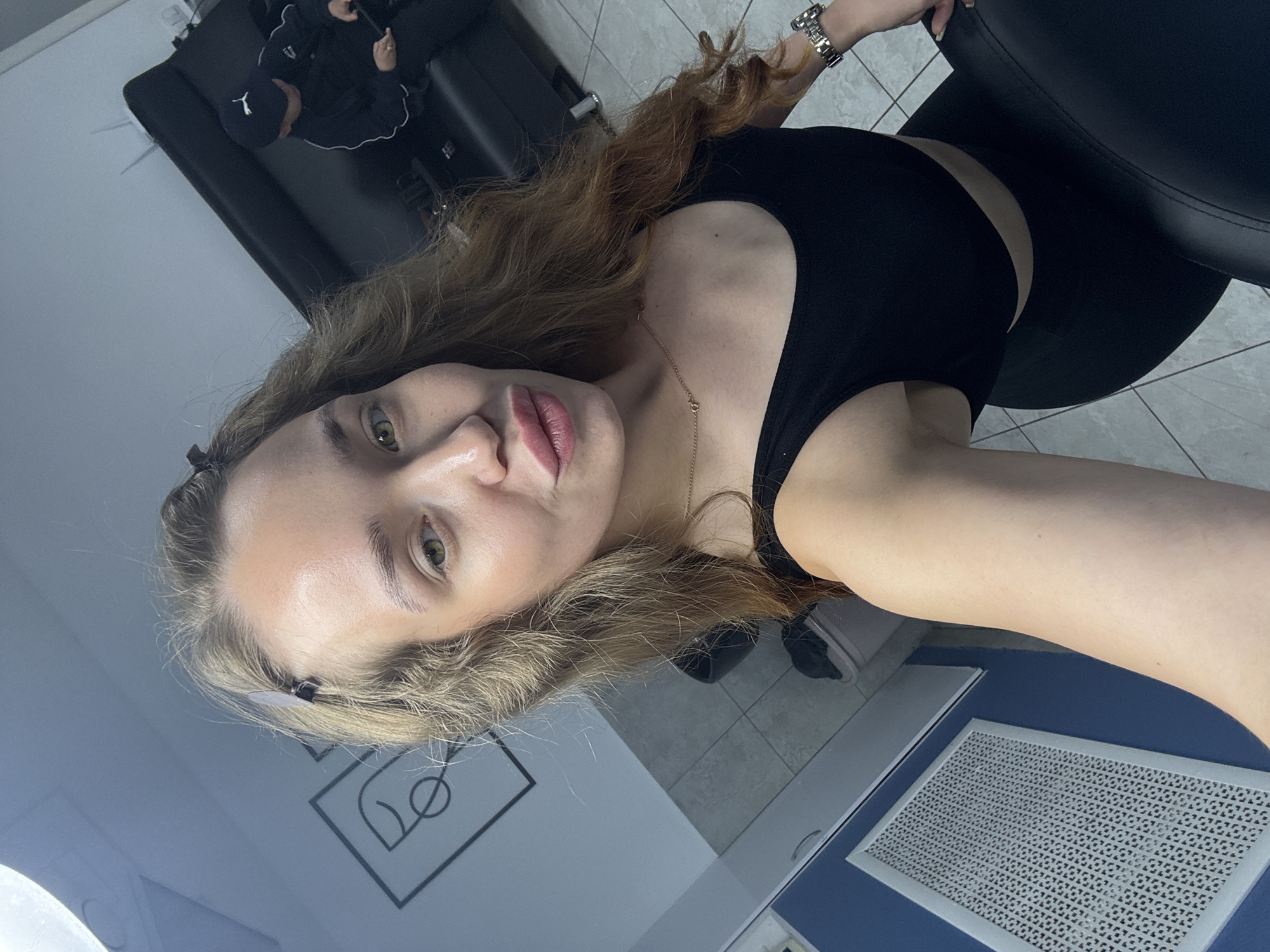 The image is a portrait of a young woman with long, wavy, reddish-brown hair and light green or hazel eyes, looking directly at the camera with a neutral expression and slightly parted lips. She is wearing a black sleeveless crop top and a delicate gold-colored necklace. A silver-colored watch is visible on her left wrist, which is positioned on the back of a black chair.
The setting appears to be indoors, possibly a salon, studio, or a waiting area. In the background, another person is seated in a black chair, wearing a dark tracksuit with white detailing and a visible "PUMA" logo on the left chest. This person also appears to be wearing a dark cap. The floor is covered with light-colored tiles.
On a blue surface in the foreground, there is a rectangular silver-colored grate with a repeating geometric pattern, potentially a ventilation cover or a tool mat. A minimalist black line graphic, consisting of interconnected circles and lines, is visible on a light-colored wall behind the main subject's head. The lighting is bright and artificial, typical of an indoor commercial space. The overall impression is a casual, selfie-style photograph.