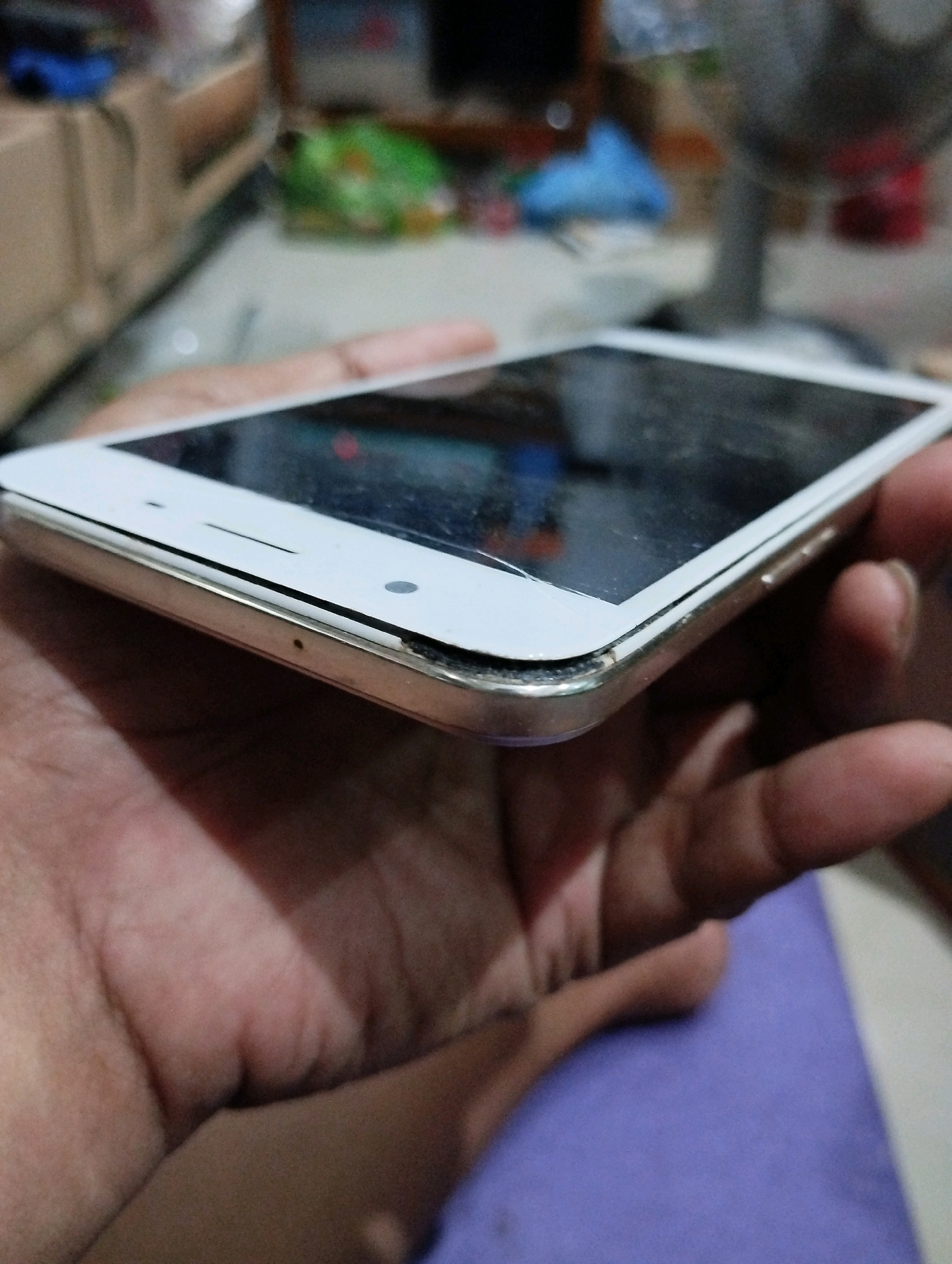 A close-up shot shows a white smartphone with a cracked screen, being held by a person's hand. The phone's metallic frame is visible, with some damage to the bezel. The background is out of focus, suggesting an indoor setting, possibly a room in Kelurahan Sidanegara, Kecamatan Cilacap, Indonesia. The lighting is dim, with some reflections on the phone's screen.