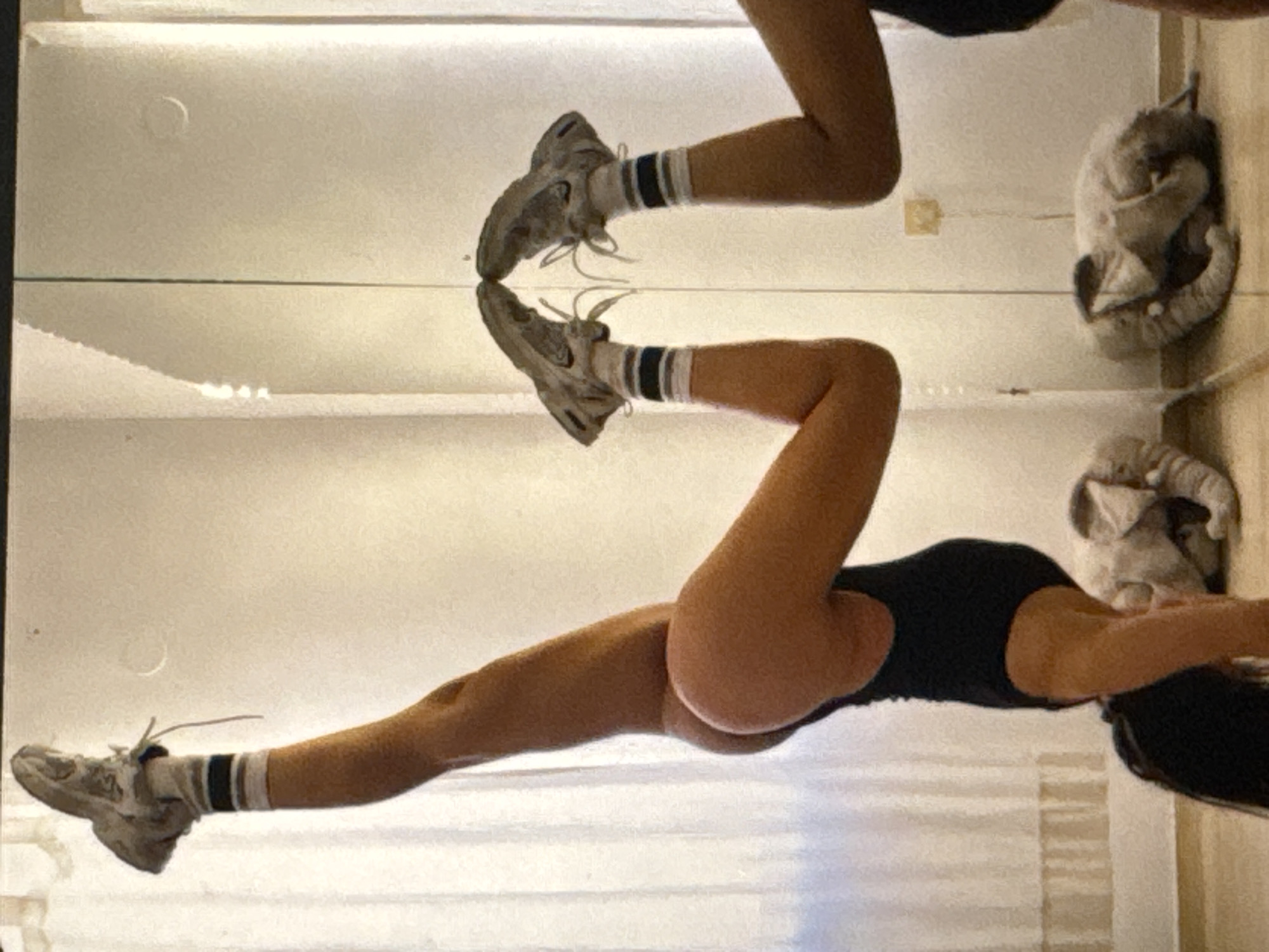 A person is performing a handstand indoors, with their body angled towards the viewer. Their left leg is extended upwards, while their right leg is bent at the knee. They are wearing a black leotard, white socks with black stripes, and athletic shoes. The person's reflection is visible in a large mirror behind them, showing a similar pose. In the background, two stuffed elephants are visible on the floor. The environment appears to be a room with white walls and curtains. This scene is located in Kuta Selatan, Indonesia.