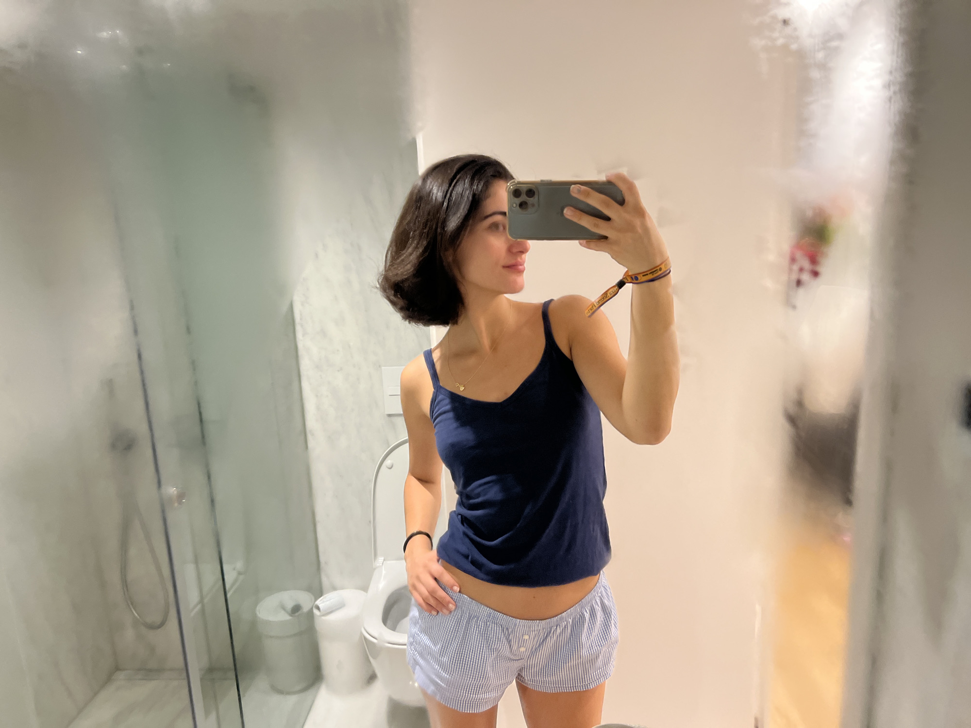 In a brightly lit bathroom, a woman with short, dark hair takes a selfie in a mirror. She is wearing a dark blue tank top and blue and white striped pajama shorts. She is holding a smartphone in her right hand, which is extended to capture her reflection. A colorful wristband is visible on her left wrist. To her left, a glass shower enclosure with a showerhead is visible. In the background, a white toilet and a trash can are present. The mirror is partially fogged, obscuring some details of the bathroom. The overall environment suggests a private dwelling, possibly a hotel room. The image does not provide any specific information indicating the location in the city of Porto, Portugal.