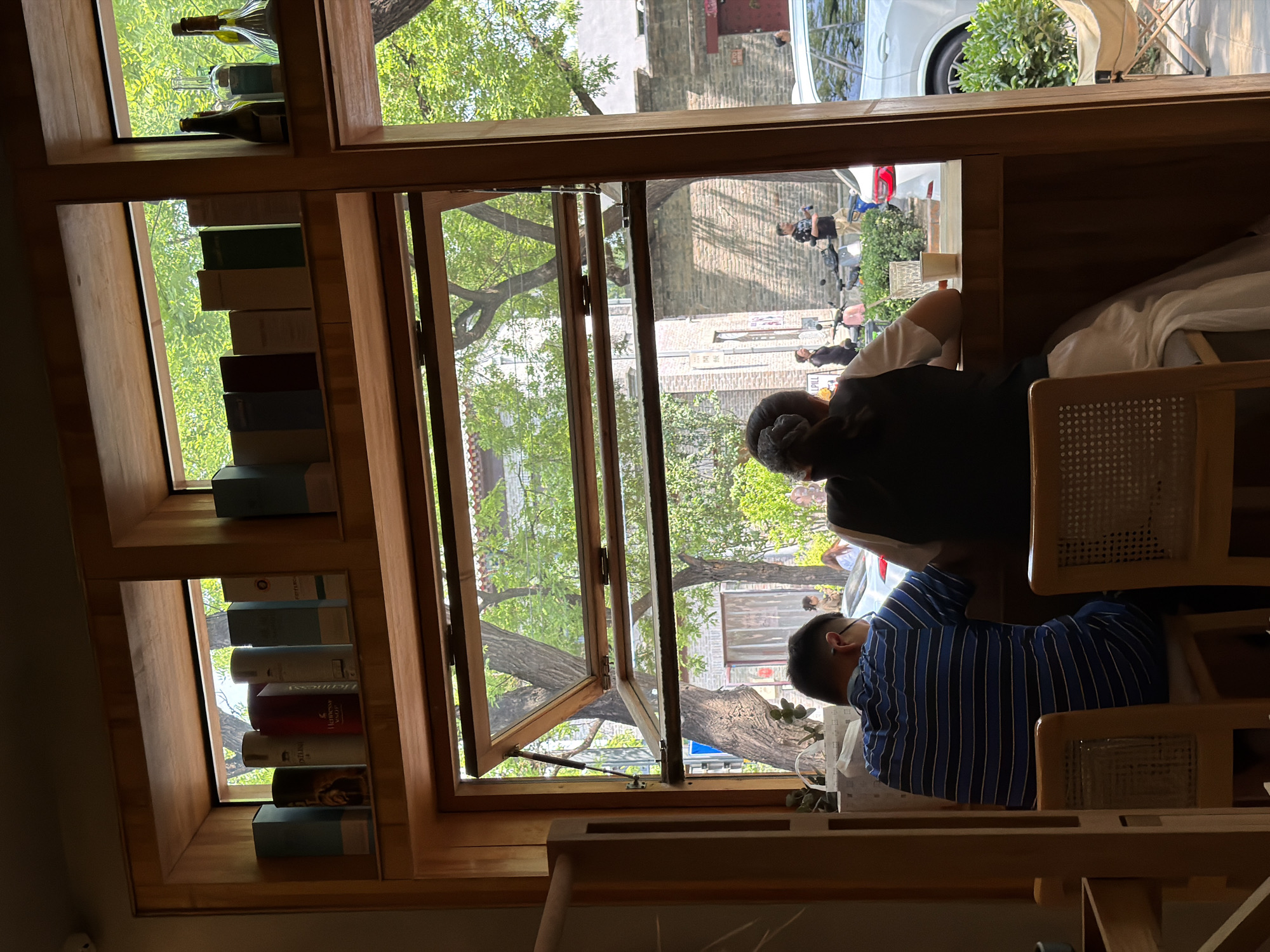 A view from inside a building, possibly a café or restaurant, shows two individuals seated near an open window with wooden frames. The location is likely in Dongcheng District, China, given the streetscape visible through the window and the prevalence of trees. The first individual, seen from behind, has dark hair tied up and is wearing a dark-colored vest or top. They are seated at a table, possibly interacting with the other person. The second individual wears a blue and white striped shirt and is also seated at the table. The window offers a view of a street outside, featuring a mix of buildings, trees, and pedestrian activity. There is a white car parked on the street, and people walking by. A shelf to the left of the window displays bottles and books.