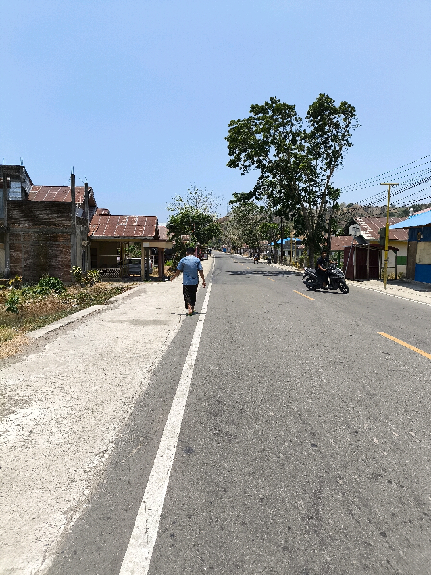 In the city of Tolongio, Indonesia, a man walks barefoot on a concrete sidewalk next to an asphalt road. He is seen from behind, wearing a blue t-shirt and dark pants. The road has a yellow line marking the center and a white line separating the sidewalk from the road. A motorcycle is driven by a man on the right side of the road. Several houses with corrugated metal roofs are visible on the left and right sides of the road, suggesting a residential or mixed-use area. A large tree with dense foliage stands prominently in the middle ground on the right. The sky is clear and blue, indicating a sunny day. The environment appears to be a rural or semi-urban area with modest buildings and vegetation.