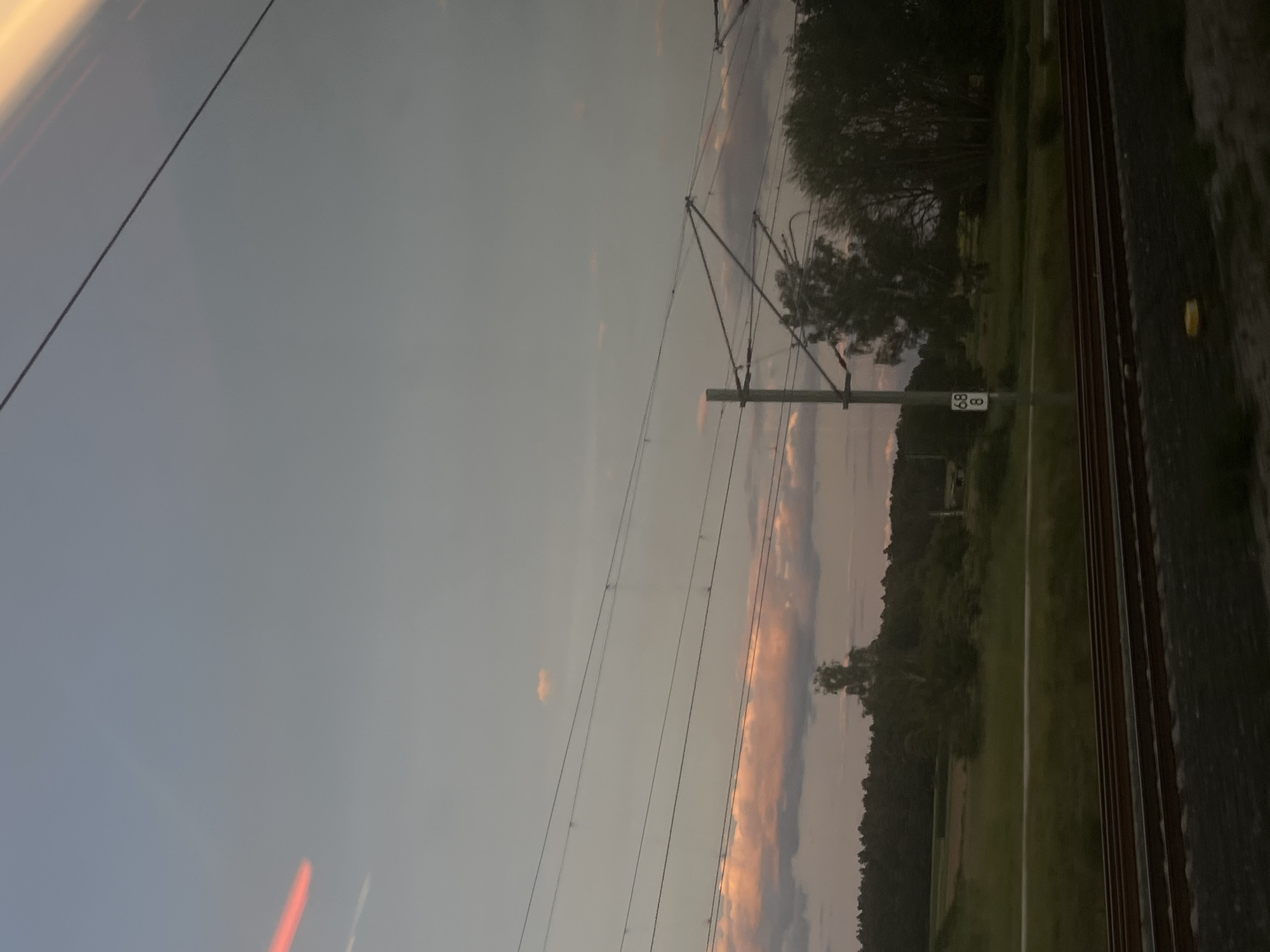 An outdoor, low-angle, eye-level shot taken from a moving vehicle shows a sky with pink and grey clouds at dusk. The sky is mostly clear with a few wispy clouds scattered throughout. Power lines crisscross the frame, supported by poles. Below the sky is a dark green tree line, with fields and a railroad track in the foreground. A signpost with the numbers "68" and "8" is visible on the left side of the frame. The image appears to be taken at dusk.