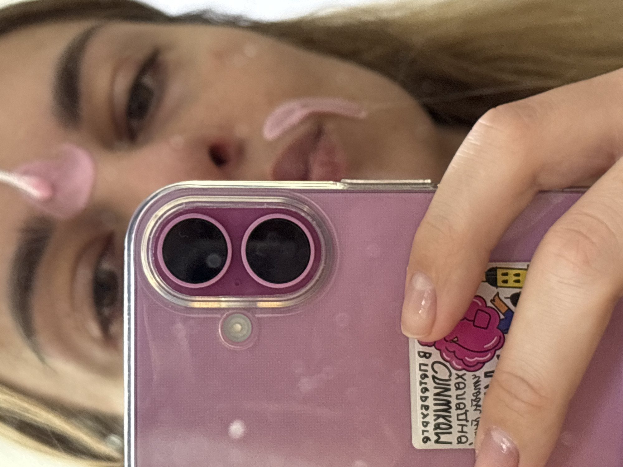 An indoor mirror selfie captures a person engaged in a skincare routine. The subject's face is visible in reflection, partially out of focus, showing one eye, an eyebrow, and parts of her nose and mouth. She has streaks of a light pink substance, likely a face mask or cream, applied to her forehead and nose area. Her light-colored hair is visible on the left side of the reflection.
The person is holding a light purple smartphone, encased in a clear protective cover, which dominates the foreground. The phone features two prominent circular camera lenses and a smaller sensor below them. Her right hand, with manicured nails (clear polish), holds the device. A sticker, partially obscured by her fingers, is affixed to the back of the phone. The text on the sticker, reversed due to the mirror reflection, reads in Russian: "Холодная СНИМКОЦ" (Kholodnaya SNIMKOTs), meaning "Cold pictures," and a sequence of numbers and letters "97892979 В".
The image portrays a personal moment of self-care, possibly taken in a bathroom or bedroom in Ust-Ilimsk, Russia. The time of day and specific weather conditions are not discernible from the image.