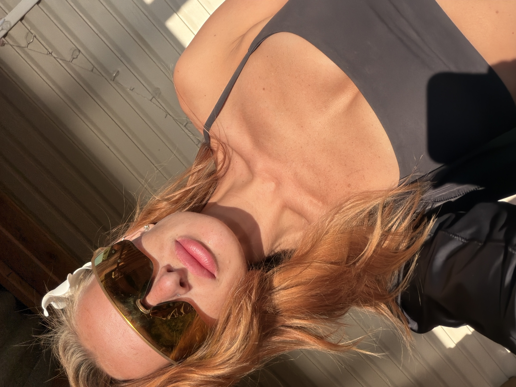A woman with red hair and sunglasses takes a selfie outdoors. She is wearing a dark grey sports bra and a black jacket. The background is a wall made of light-colored horizontal planks, with some sunlight casting shadows. There is a white flower decoration in her hair. She is wearing a nose ring and earrings. The image is taken at an upward angle, with the woman's face and upper body prominently featured.