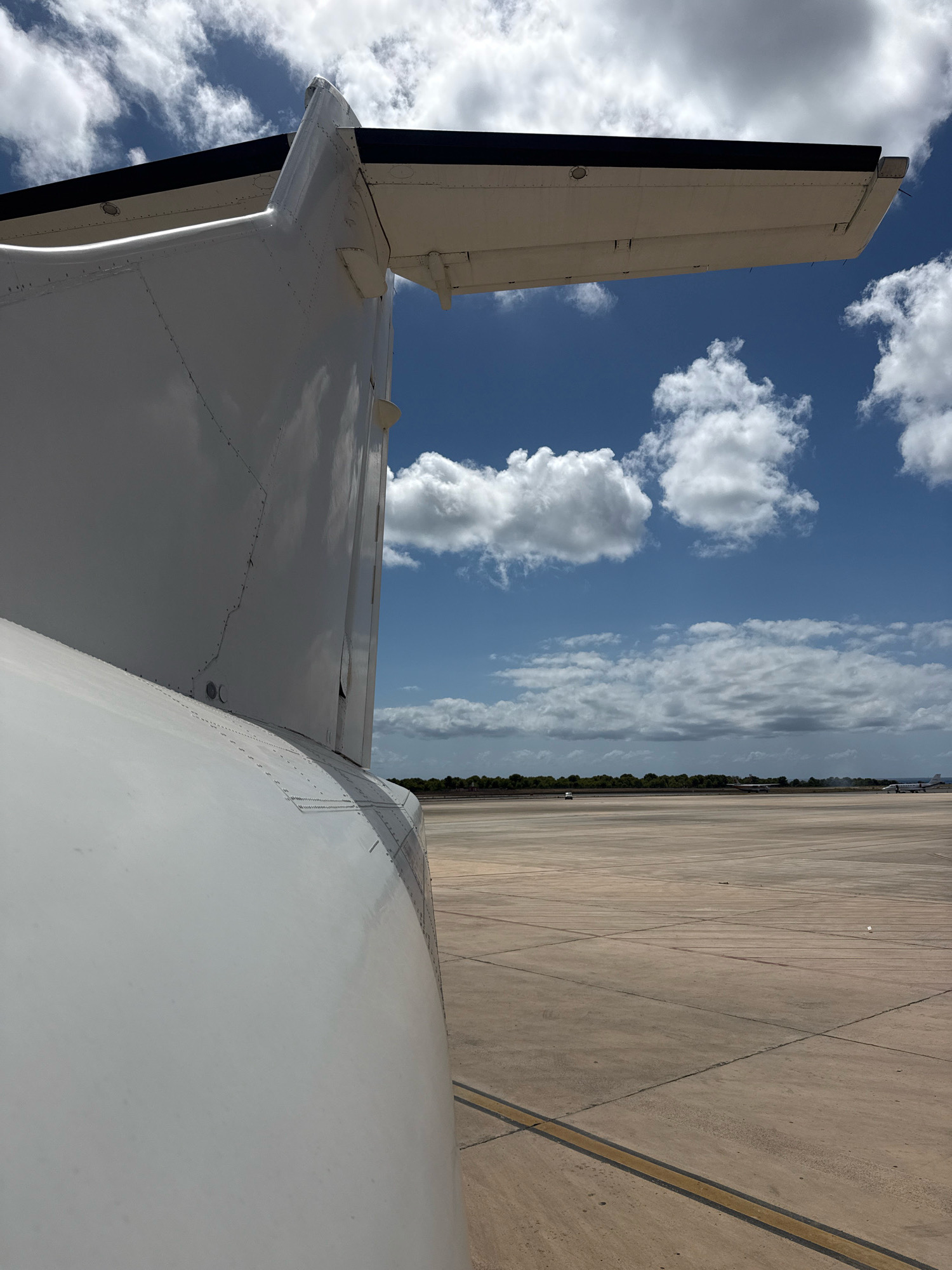 This is a close-up, low-angle shot of the tail section and a portion of the wing of a white airplane, positioned on an airport tarmac under a bright, cloudy blue sky. The airplane's tail is in the foreground, dominating the left side of the frame and curving away from the viewer. Its white surface reflects the sunlight, showing rivets and panel lines. The wing, also white with a dark leading edge, extends horizontally towards the right, partially obscuring the sky.
The background depicts a wide, concrete airport runway stretching into the distance. Several small aircraft are visible on the tarmac in the mid-ground, appearing as distant specks. A line of green trees forms a horizon line behind the runway, and beyond that, a hazy landscape suggests the presence of a distant town or city. The sky is filled with scattered white cumulus clouds against a vibrant blue backdrop. The overall impression is a sunny day at an airport, with a focus on the aircraft's structure.
The image is likely taken at Ibiza Airport (IBZ), located near the town of Sant Josep de sa Talaia, Spain. This is a reasonable assumption given the characteristic landscape and the presence of a significant airport.