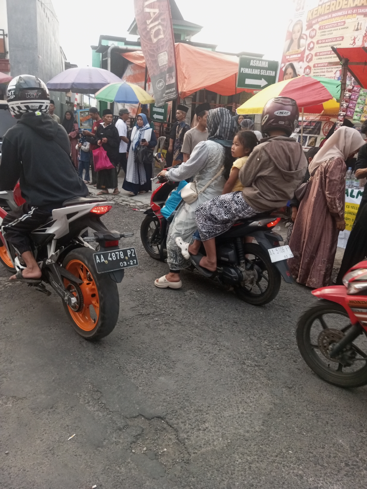 In Pandansari, Indonesia, a street scene unfolds with several individuals on motorcycles. In the foreground, a person wearing a black helmet and hoodie is on a white and red motorcycle, facing away from the camera. To their right, another motorcycle carries a rider in a grey jacket and a passenger wearing a light blue hijab and patterned skirt, with a young child seated in front of them. Further back, people in traditional Islamic attire are visible, some carrying shopping bags. A sign displaying "ASRAMA PEMALANG SELATAN" with an arrow points to the left, suggesting a location or destination. The road surface is asphalt and appears somewhat worn. Several colorful umbrellas and awnings are visible in the background, along with buildings and banners with text, one of which prominently displays "MERDEKA". The overall atmosphere suggests a busy, perhaps market or event-related, outdoor gathering.