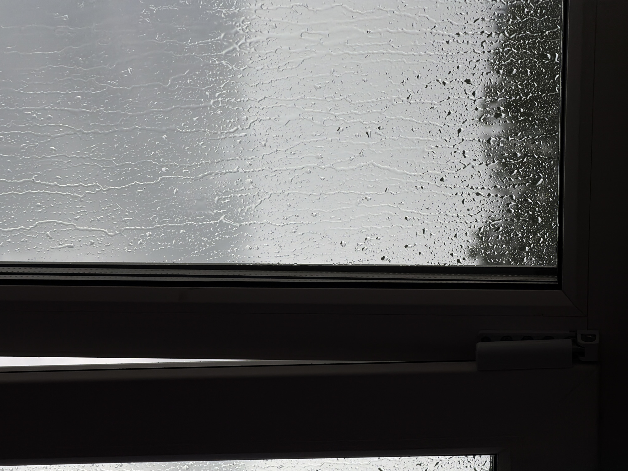 The image shows a close-up view of a window with raindrops on it, obscuring the view of the outdoors. The window frame is white, and on the left side of the frame, a darker, possibly closed shutter or curtain is visible. The right pane of the window is covered in streaks and droplets of water, with a faint, blurry outline of trees visible in the distance. The sky appears overcast and gray. The overall scene evokes a rainy day. The location is identified as Ust-Ilimsk, Russia. There are no people or discernible actions captured in the image.