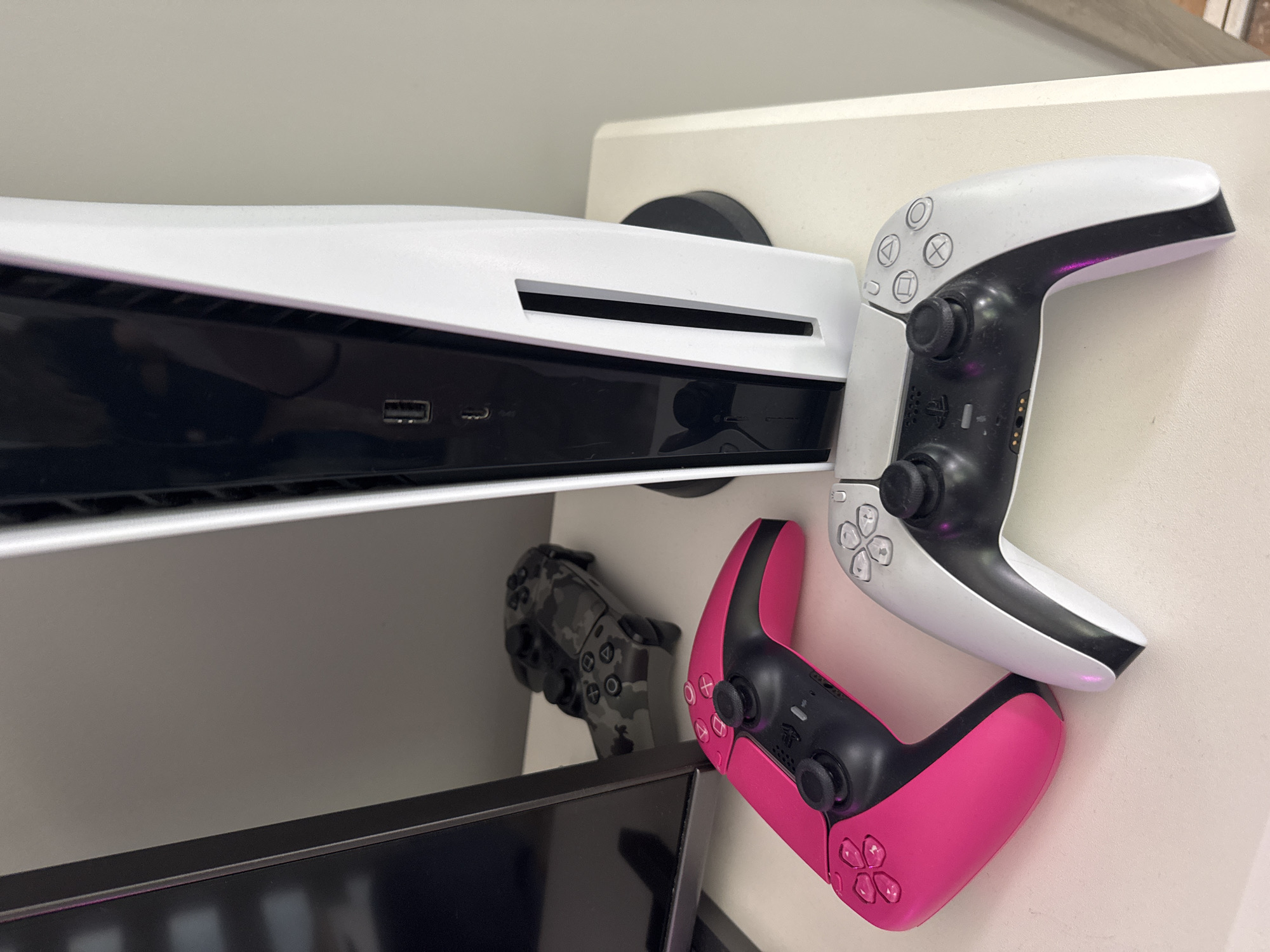 The image displays a modern gaming setup in an indoor setting. A white PlayStation 5 console stands vertically on a black circular base, positioned on a light-colored surface, likely a desk or shelf. The console's front panel is visible, featuring a USB-A port and a USB-C port, along with a vertical ventilation slot. To the left of the console, a vibrant pink PlayStation 5 DualSense controller rests on the same surface. To the right, partially beneath the console, is a standard white and black PlayStation 5 DualSense controller. In the background, behind a black monitor or television screen partially visible on the left, a third PlayStation 5 DualSense controller with a camouflage pattern is present. The background consists of a plain, light-colored wall. No people are visible.