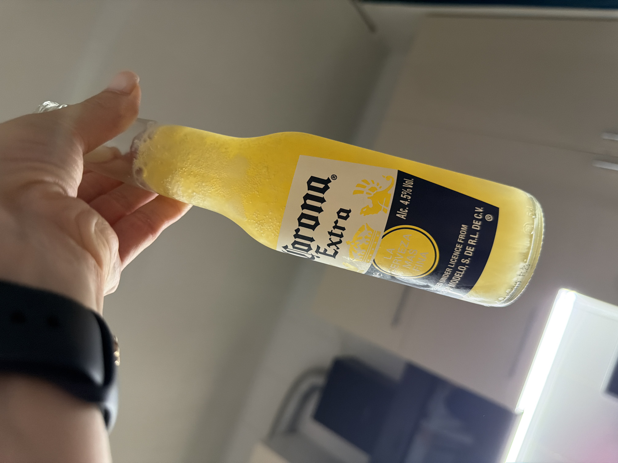 A close-up, low-angle shot shows a hand holding a frosty bottle of Corona Extra beer upside down. The bottle is filled with yellow liquid and condensation droplets cover its surface. The label on the bottle is visible, displaying the Corona Extra logo, a crown, and the text "LA CERVEZA MAS FINA" and "Alc. 4.5% Vol.". The hand is wearing a black smartwatch on the left wrist and a ring on the index finger. The background is blurred, showing indistinct kitchen cabinetry and a bright light fixture. The scene appears to be indoors.