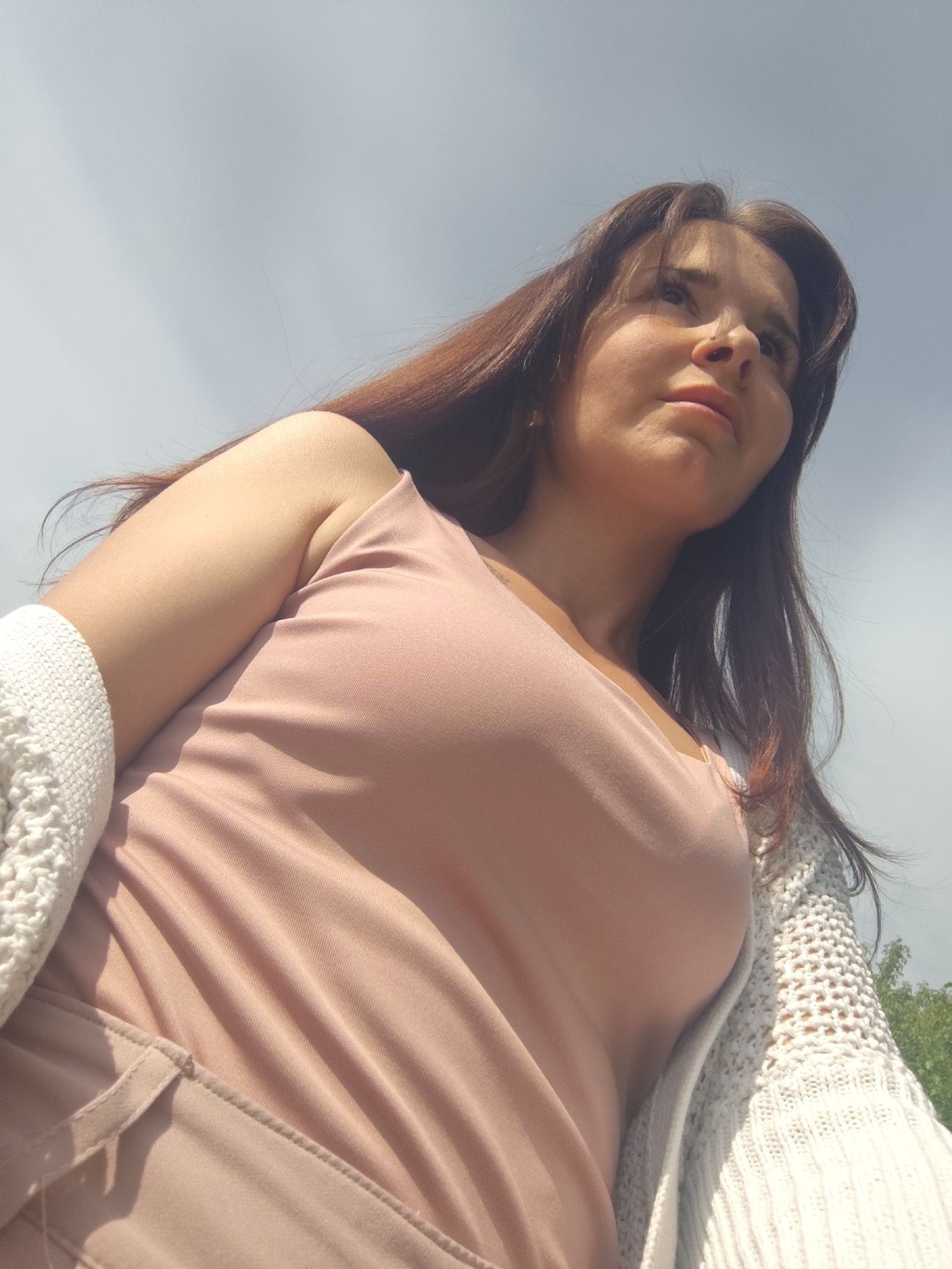 A young woman is viewed from a low angle, looking upwards and slightly to the right. She has long, dark hair and is wearing a light pink top. A white, crocheted cardigan is draped over her left shoulder. The background is a cloudy sky. The location cannot be determined from the image.