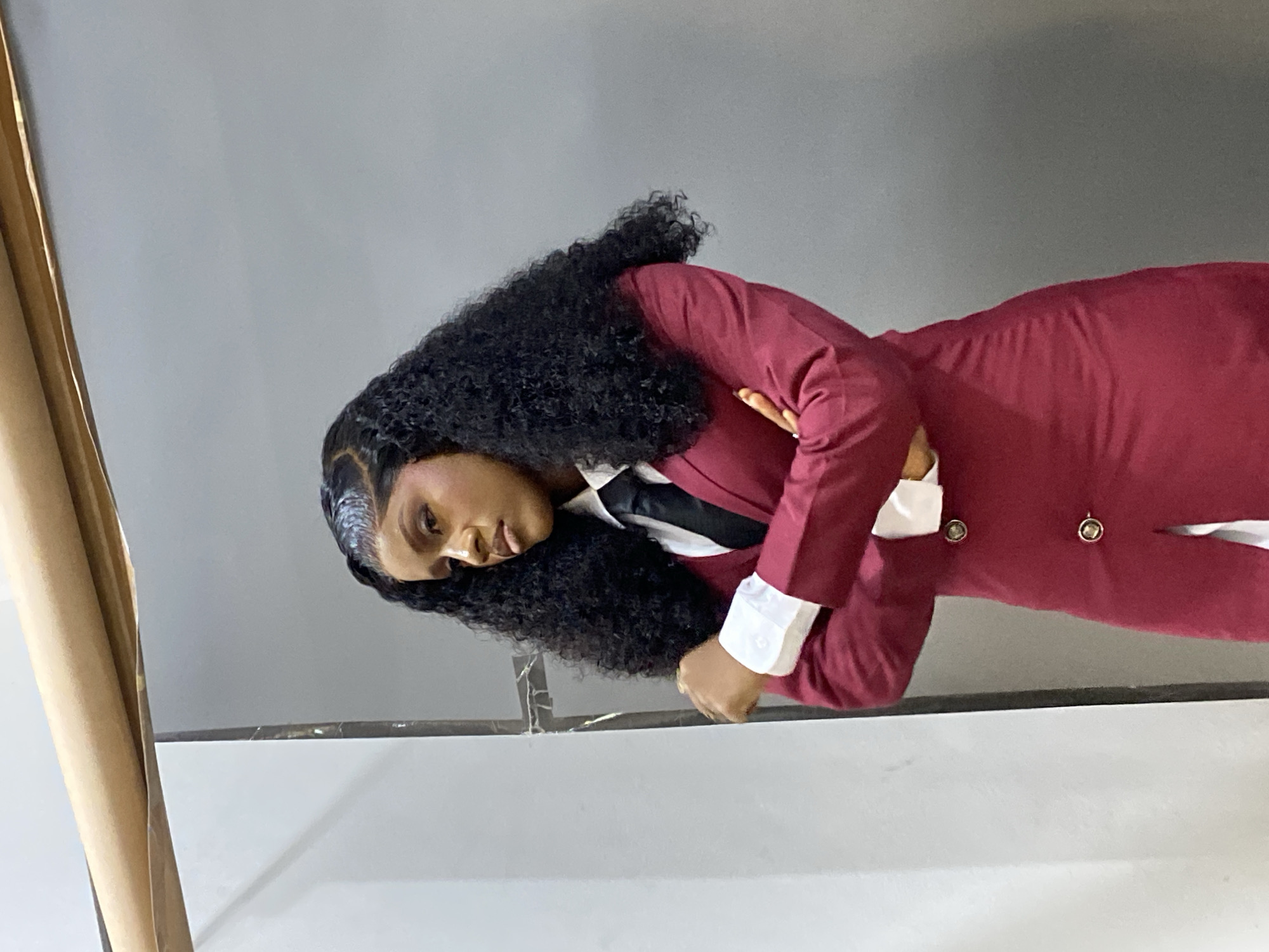 A young Black female stands in an indoor setting in Gura Top, Nigeria, with her arms crossed over her midsection. She is dressed in a white collared shirt, a black tie, and a maroon blazer-style dress or long blazer with two visible gold buttons on the front. Her long, dark, curly hair is styled with a defined hairline and she wears makeup including eyeliner and possibly lip gloss. Her expression is serious or contemplative as she looks slightly towards her left. The background features a split wall with a light grey or white panel on the left and a darker grey section on the right, separated by a thin, dark vertical structure. At the top of the frame, a beige rolled object rests on a horizontal support. The scene is evenly lit.