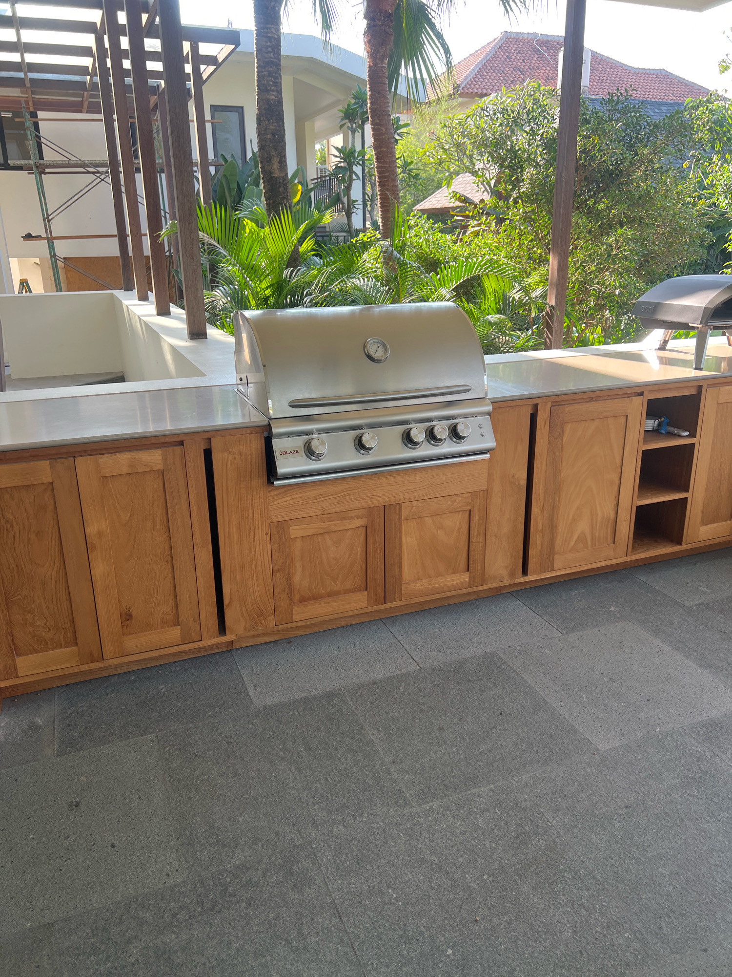 The image depicts an outdoor kitchen area located in Ngaglik, Indonesia. The focal point is a built-in stainless steel barbecue grill, labeled "UBLAZE," positioned on a light-colored countertop. Beneath the countertop, a series of wooden cabinets with plank-style doors provide storage. To the right of the grill, a smaller black pizza oven is visible. The kitchen area is situated on a patio paved with large, dark grey rectangular tiles. In the background, lush tropical foliage, including palm trees and other dense greenery, creates a vibrant natural setting. Behind the foliage, a building with a red tiled roof and a white facade is partially visible. The overall scene suggests a modern outdoor living space, likely in a residential or hospitality context, with elements of both functional design and integration with the natural environment.
