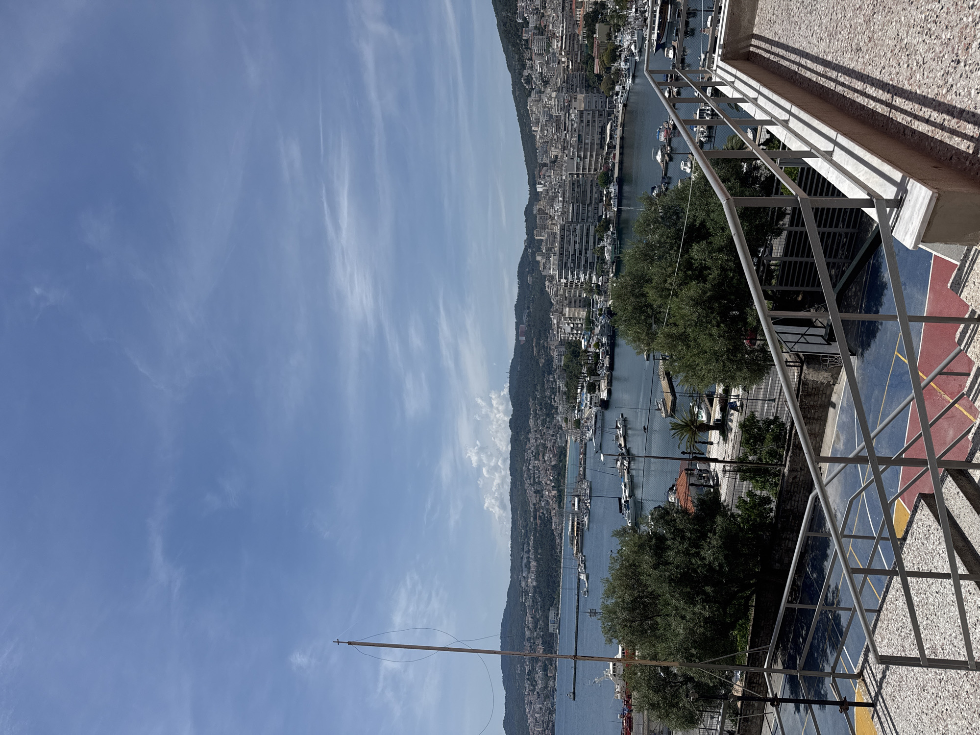 This image captures a panoramic view of the city of Kavala, Greece, featuring a harbor filled with numerous boats and a cityscape climbing the hills. In the foreground, a portion of an outdoor basketball court with blue and red surfaces is visible, bordered by a gray railing and textured stairs. Beyond the court, lush green trees partially obscure the view of the harbor. The harbor itself is a hub of activity, with a dense collection of various boats, including sailboats and motor yachts, docked in the clear blue water. A long, slender pole with a cross at its apex stands prominently in the left side of the frame. The backdrop is a sprawling urban landscape of white and light-colored buildings that extend up the gentle slopes of the surrounding hills, dotted with more greenery. The sky above is a vibrant blue, streaked with wispy white clouds, indicating fair weather. The overall scene suggests a sunny day in a coastal Mediterranean city.