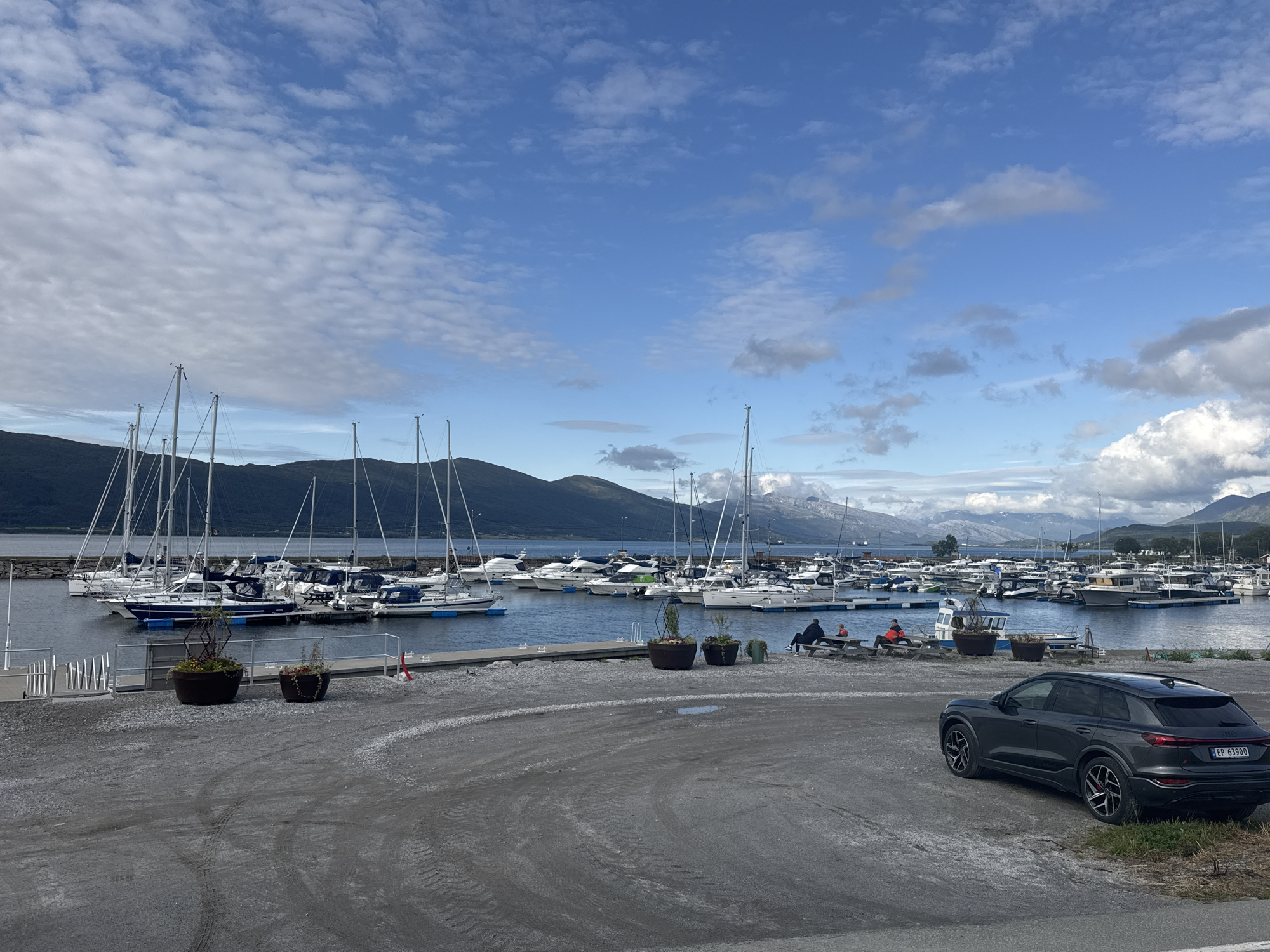This media file depicts a scenic view of a marina in Nesna, Norway. The sky is a vibrant blue, dotted with fluffy white clouds, suggesting pleasant weather, possibly late afternoon. The marina is filled with numerous sailboats and motorboats docked in the calm, dark blue water. In the background, majestic, verdant mountains rise, some capped with snow, adding to the dramatic natural beauty of the location.
In the foreground, a gravel area leads to the marina. A dark gray SUV is parked on the right, with a license plate clearly visible reading "EP 63900". To the left of the SUV, two large planters with small trees and plants add a touch of greenery. Further left, a couple of people are seated at a picnic table, seemingly enjoying the view. Another person is seated on a bench. The overall atmosphere is peaceful and serene, indicative of a relaxed day at the harbor. The presence of boats and the mountainous backdrop strongly suggest a coastal or fjord setting.