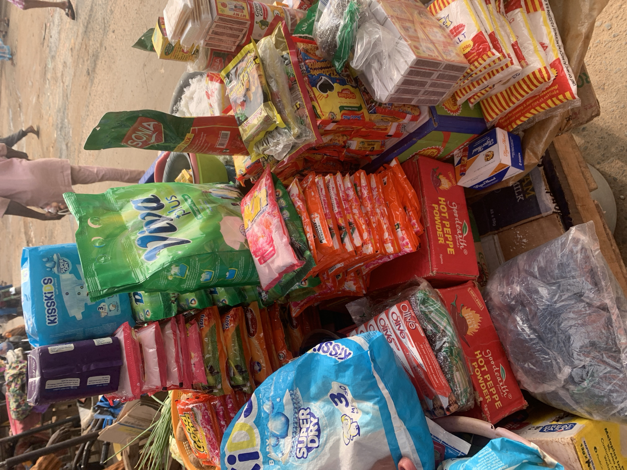 This image captures a vibrant market scene in Shagamu, Nigeria, likely during daylight hours. The focus is on a colorful assortment of goods piled high, suggesting a vendor's stall. Dominating the foreground are various packaged items, including what appear to be diapers, detergent powder, seasoning packets, and other household essentials. Brightly colored packaging in shades of blue, green, orange, and red creates a lively visual.
Text visible on the packaging includes brand names like "KISSKIS," "SONIA," "Mamuda," "Olive," and "Tiger Head." There are also product descriptions such as "BABY DIAPERS," "SUPER DRY," "Detergent Powder," "HOT PEPPE POWDER," "SALT," and "AM MILK." Some text indicates weight and product quantity, like "250g" and "5gms."
In the background, out of focus, are glimpses of people, including a person wearing a pink top and grey pants. Their presence suggests the setting is a bustling public place where commerce is taking place. The ground appears to be a dusty, unpaved surface, common in market areas. The overall impression is one of a lively and active marketplace, showcasing the variety of products available to shoppers.