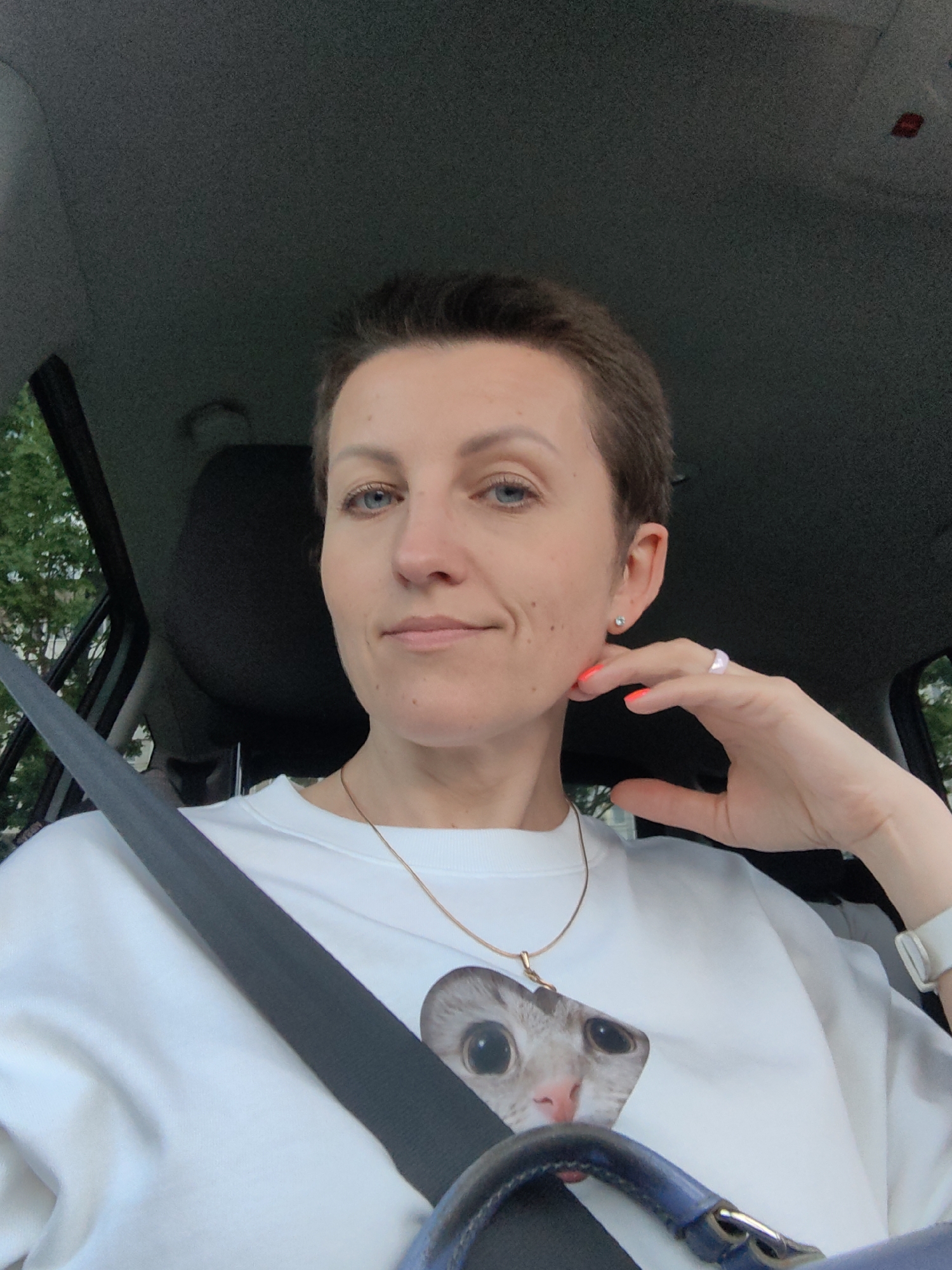 The image shows a medium shot of a woman in a car, likely a passenger, with a seatbelt across her chest. She has short, brown hair, blue eyes, and fair skin. She is wearing a white sweatshirt with a graphic of a cat's face on it, a thin gold chain necklace, a stud earring in her left ear, and a large, light-pink ring on her right hand, which she is resting on her chin. Her fingernails are painted a bright orange. The background consists of the car's interior, including the headrest and a portion of the dashboard. Trees and greenery are visible through the car window on the left. The overall lighting suggests it is daytime. The image does not provide any specific visual cues that would indicate the location is Minsk, Belarus.