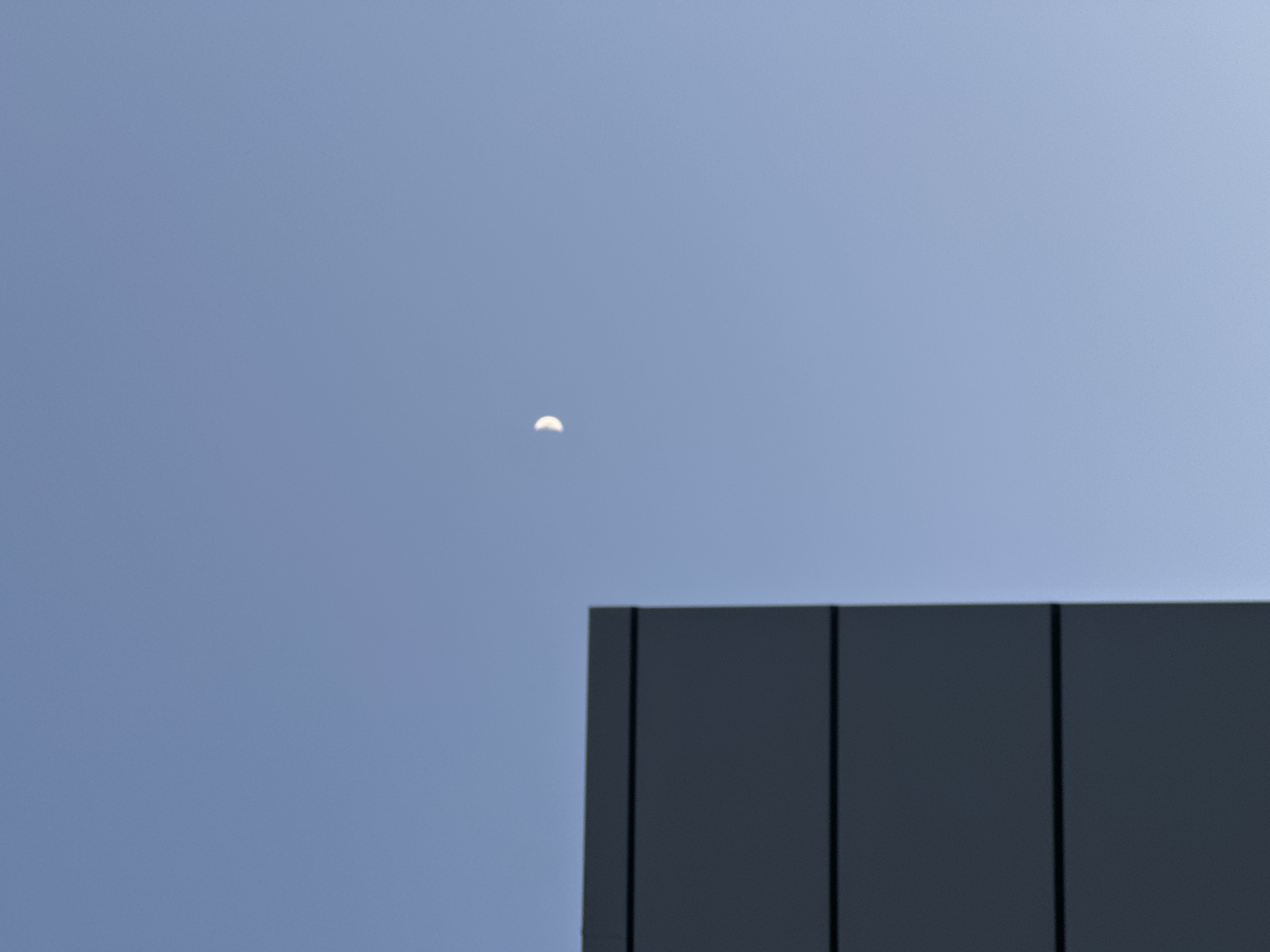 The image shows a crescent moon in a light blue sky above a dark gray building. The building appears to have a panelled exterior with vertical lines separating the panels. The location is assumed to be Chaikovsky, Russia. No people, actions, or interactions are visible.
