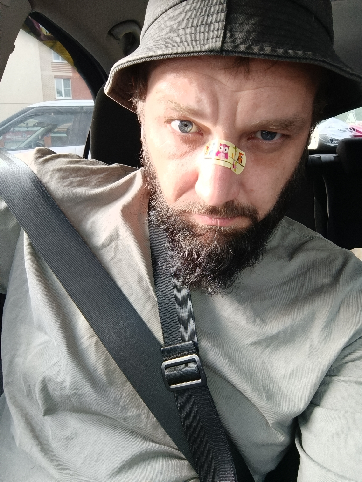 A man wearing a bucket hat and a light-colored shirt sits in the driver's seat of a car, his seatbelt fastened. He has a beard and a band-aid with owl illustrations on the bridge of his nose. The man is looking towards the camera with a serious expression. The background shows a building and another car through the car window, suggesting an urban or suburban environment. The lighting indicates it is daytime. There are no explicit indicators of the location being Arkhangelsk, Russia.