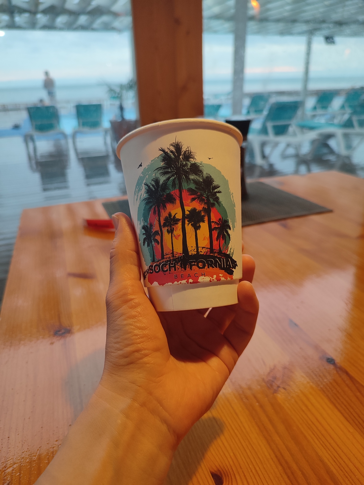 A person is holding a paper cup with a "Sochifornia Beach" design featuring palm trees and a sunset, inside a building with a view of the coastline in Sirius, Russia. Through the windows, beach chairs and a blurred figure of a person can be seen. The cup is held over a wooden table. The lighting is ambient and slightly subdued.