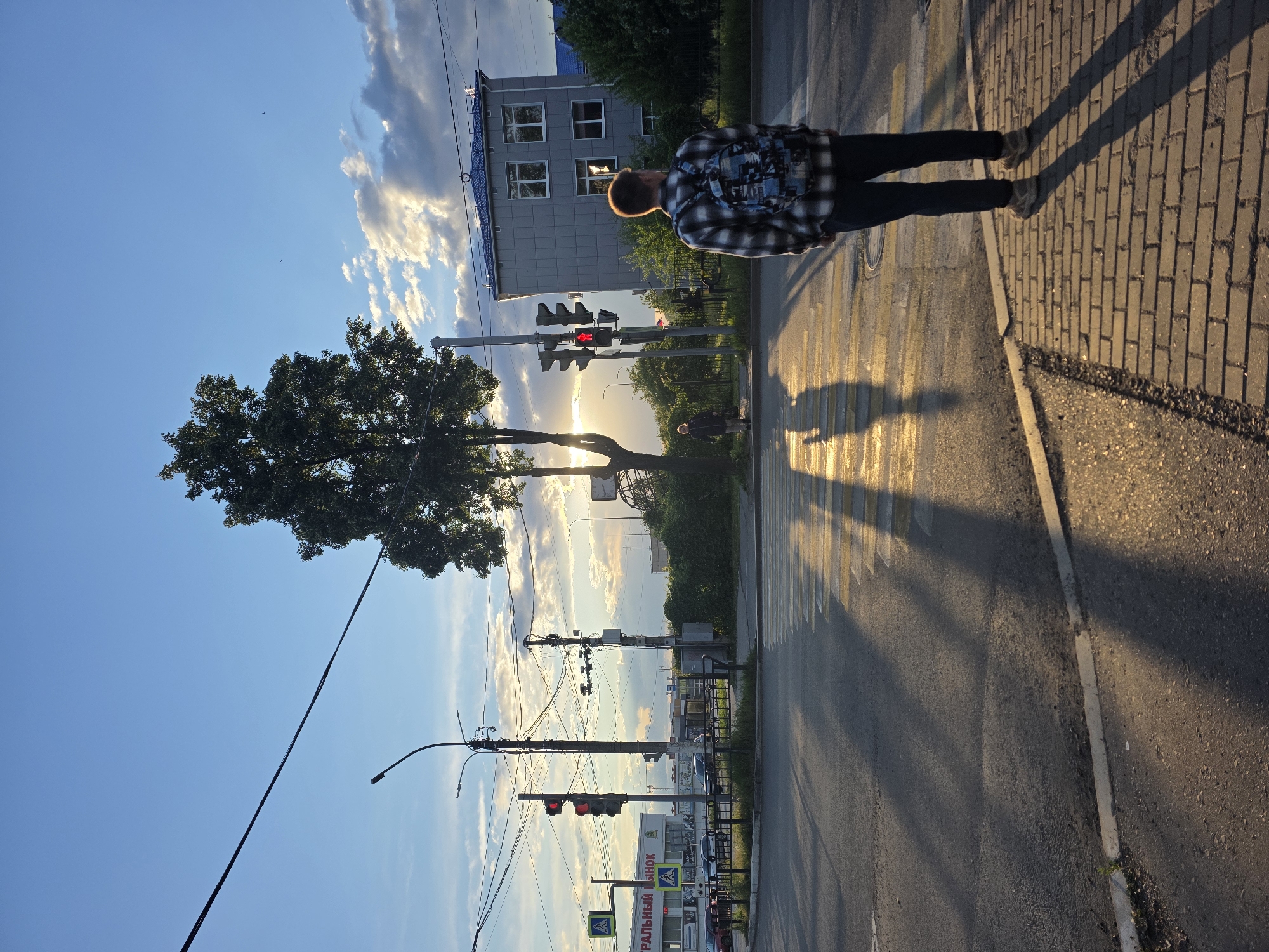 A photograph captures a man standing on a pedestrian crossing in Chaikovsky, Russia, during what appears to be late afternoon or early evening, given the long shadows and setting sun. The man, wearing a plaid shirt and dark trousers, is walking towards the sidewalk lined with brick pavers. A traffic light shows a red signal. Sunlight filters through a tree and street fixtures, casting pronounced shadows across the crosswalk. A multi-story building is visible in the background, along with utility poles and wires stretching across the sky. A store front bearing the name "ФИРАЛЬНЫЙ РЫНОК" can be seen in the distance.