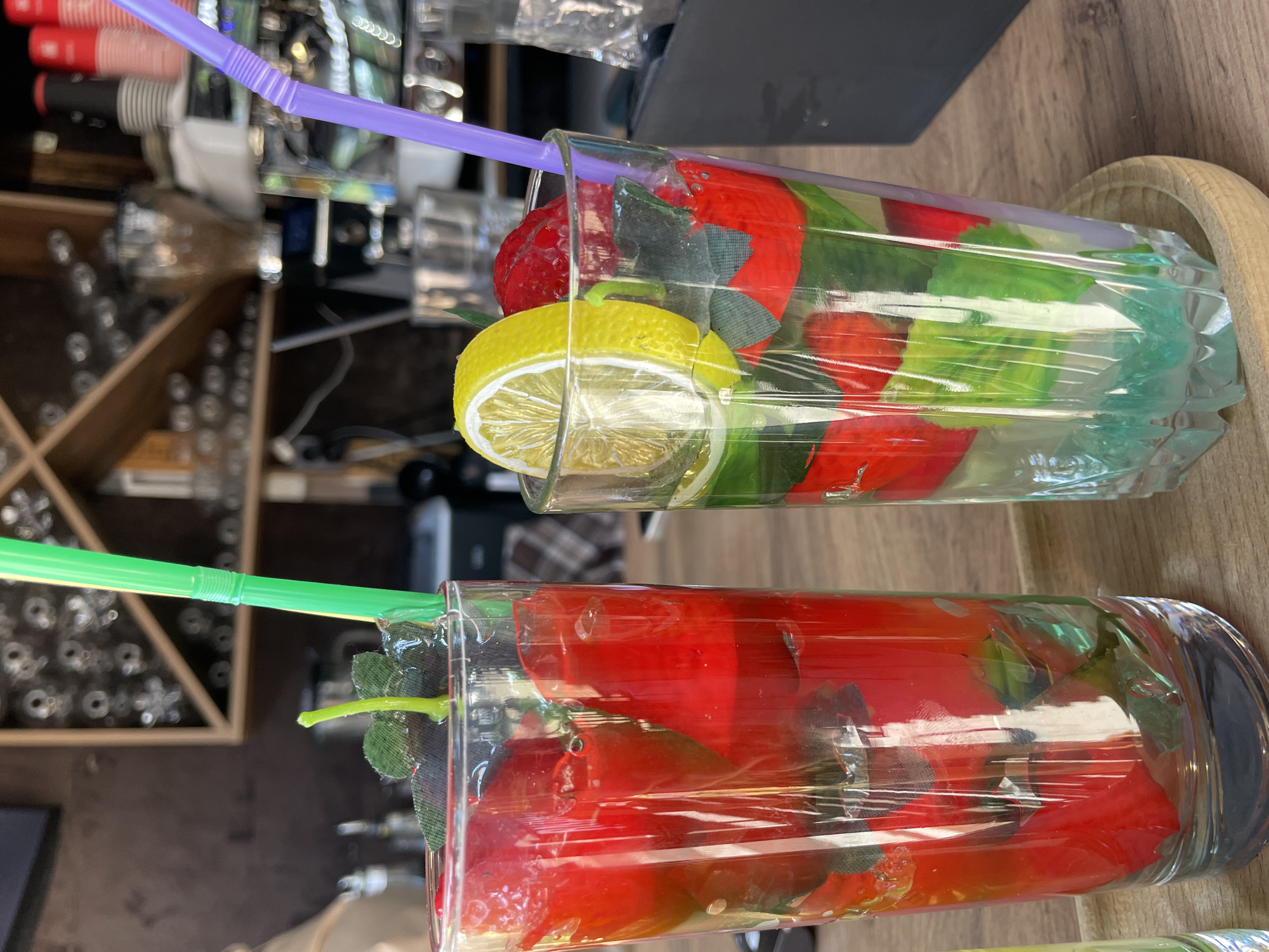 Two tall glasses filled with red and green elements, likely artificial fruits and leaves, sit on a wooden surface. The glass on the left contains primarily red pieces and a green straw. The glass on the right has a slice of lemon on top, red and green elements within, and a purple straw. The background shows shelves with various items, possibly in a cafe or bar setting. The environment appears to be indoors, with a focus on the drinks. The image does not provide any specific details about the city of Sochi, Russia, or any people.