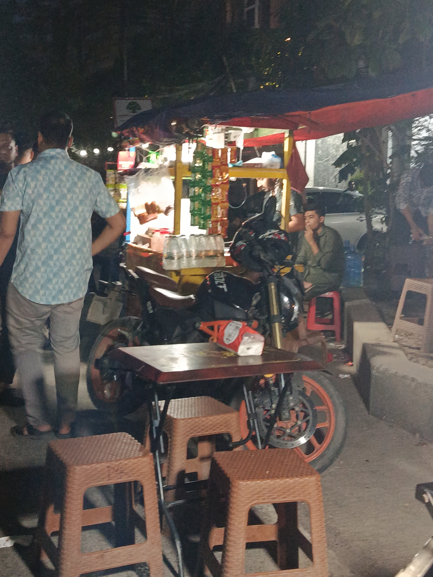 The image shows a street scene in Dhaka, Bangladesh, at night. A food stall is set up with a yellow cart and red and blue tarps overhead. A motorcycle is parked in front of the stall. There is a table with four brown plastic stools in the foreground. A man in a green shirt sits on a red stool near the cart, smoking. Other people stand nearby, including a man in a blue patterned shirt. Bottles of water are on the ground near the seated man. The environment is dimly lit.