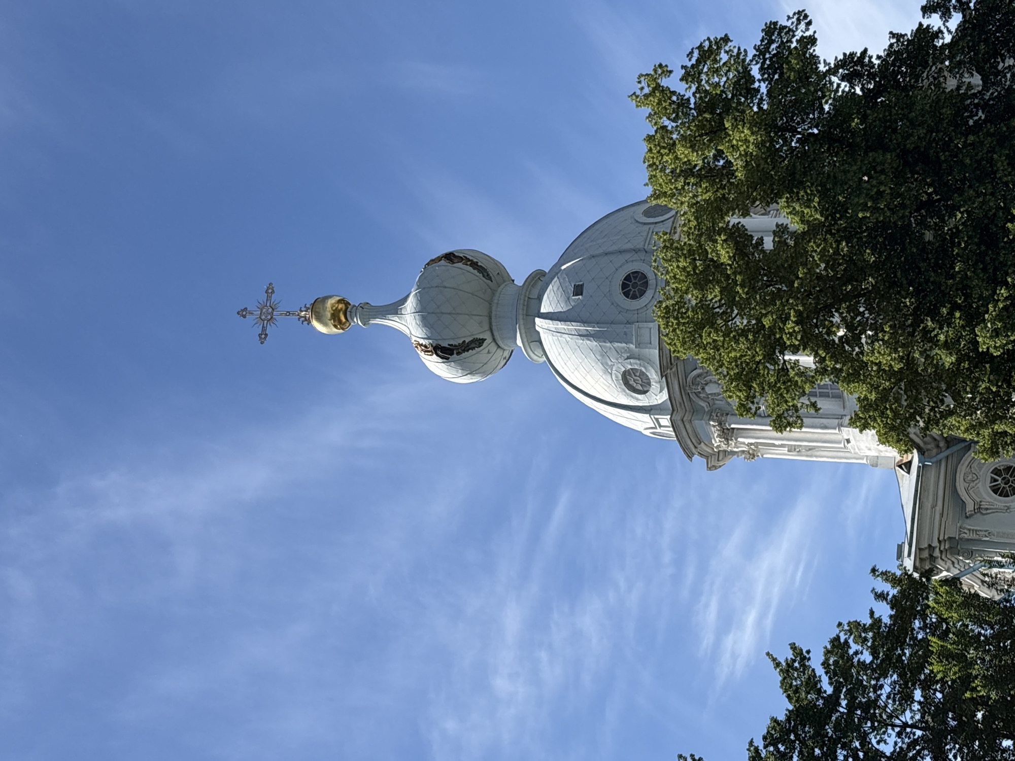 This image shows the dome and spire of a building, likely a church or cathedral, against a bright blue sky. The dome is white with intricate patterns and circular windows. A golden cross adorns the top of the spire. Lush green trees frame the building on the right and at the bottom of the image. The scene is well-lit, suggesting it's daytime, and the clear sky with wispy clouds indicates fair weather. The architectural style points towards a European setting, and the provided context confirms it is Saint Petersburg, Russia. There are no people or visible text in the image. The perspective is from below, looking up at the structure, which gives a sense of grandeur and height. The overall impression is one of architectural beauty and a peaceful, clear day.