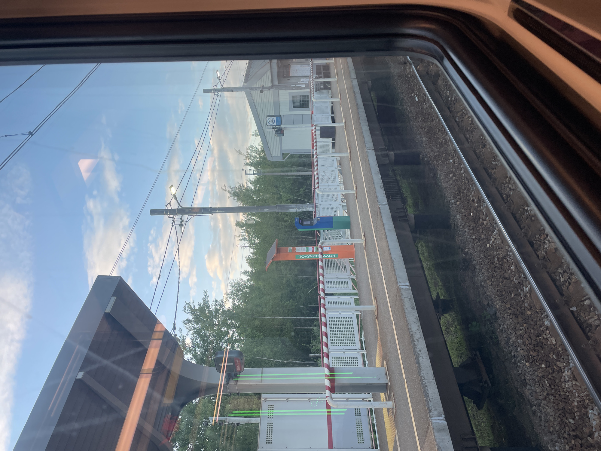 A view from inside a train looks out a window at a railway station platform under a bright, partly cloudy sky. The image is oriented so the platform runs horizontally across the frame, with train tracks in the immediate foreground and dense green trees forming the background.
On the platform, several white and red striped barriers are visible, delineating the edge. A small, light-grey station building with a dark roof and a prominent window stands behind these barriers. A blue and white sign, possibly a station or railway logo, is affixed to the building. Further along the platform, an orange stand features the Cyrillic text "ПОЛУЧИТЕ ТАЛОН," which translates to "GET A TICKET" or "GET A TOKEN/VOUCHER." Nearby, a modern grey structure with glowing green light lines and blue and green waste bins are also present. Several utility poles with overhead wires crisscross the sky, indicating an electrified railway line.
The train appears to be stationary or moving slowly past the platform. No people are visible, and the scene depicts a quiet moment at the station during daylight hours with clear weather. Reflections from the train's interior are visible on the window glass. The overall impression is a factual depiction of a railway stop in Gribanovo, Russia.