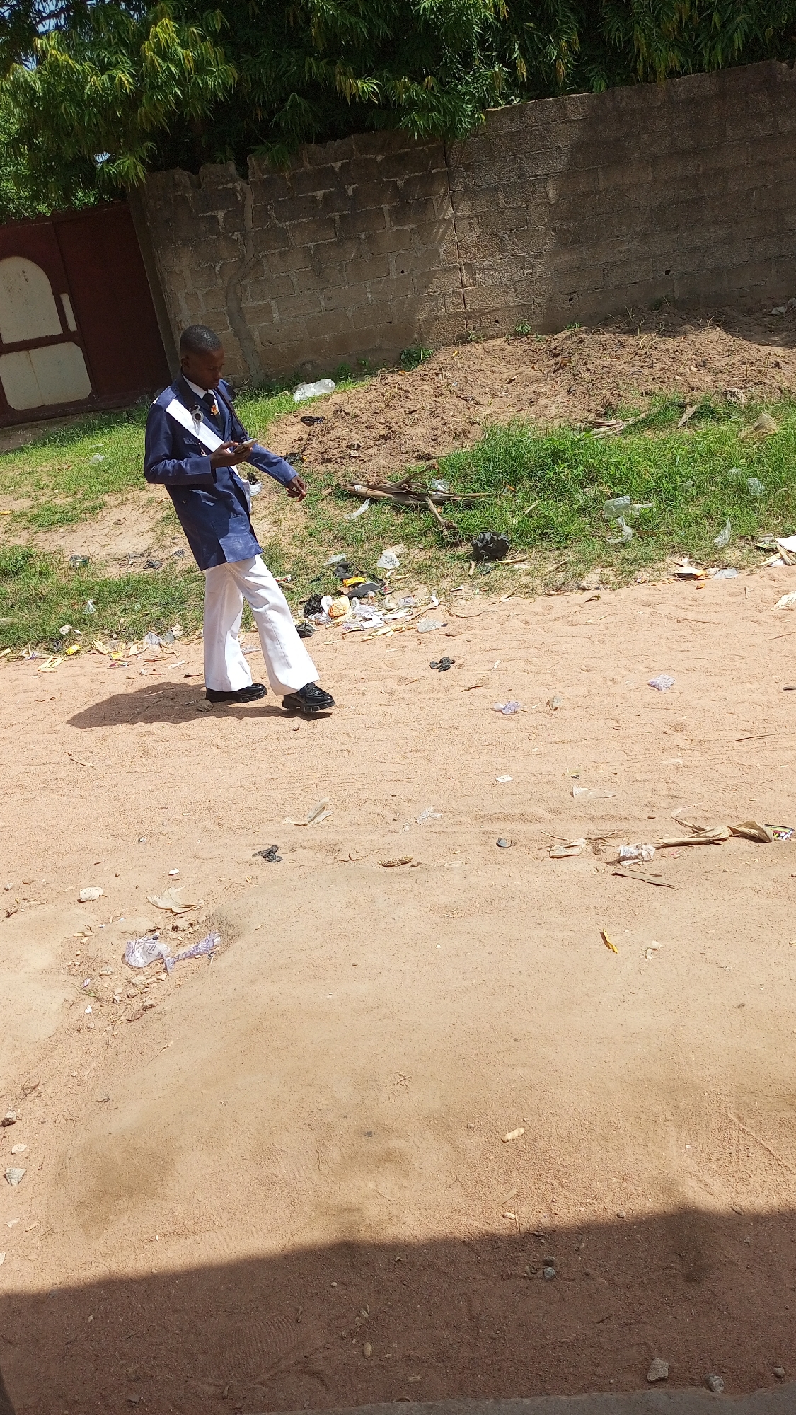 A young Black male, likely a student, stands on a dirt and sand path in Jalingo, Nigeria. He is wearing a dark blue blazer with white trim and white flared trousers, with black shoes. His right hand holds a smartphone, and his left hand is extended as if gesturing or about to reach for something. The environment is dusty, with patches of dry grass and scattered litter, including plastic bottles and paper debris, along the path and the grassy verge. Behind him, a low, rough brick wall with vertical support pillars forms a backdrop. Overgrown green foliage is visible above the wall. A large, weathered, reddish-brown metal gate with white panels is situated to the left of the frame, near the wall. The ground in the foreground is uneven, with a significant mound of dirt on the left.