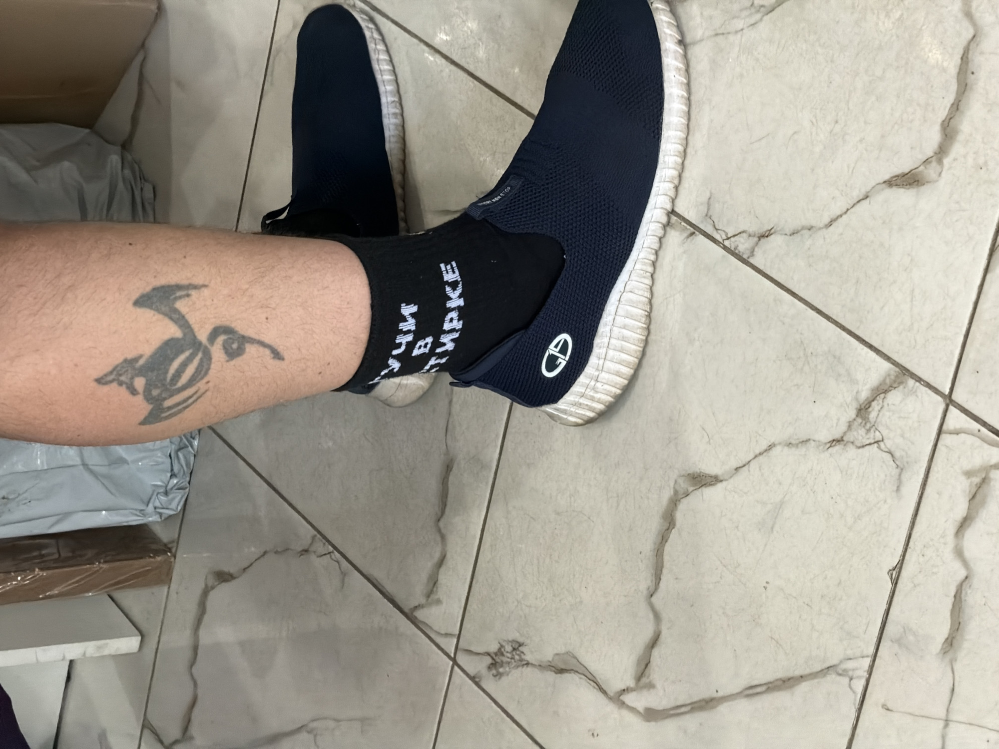 The scene is located indoors in Saint Petersburg, Russia, as indicated by the Cyrillic text on the visible sock.
The image captures a low-angle view of a person's lower right leg and foot, positioned on a light-colored tiled floor. The skin on the leg is Caucasian with visible hair. A black, stylized tattoo, resembling an abstract creature or mythical beast, is visible on the upper calf/shin area.
The person is wearing a black ankle-length sock with white Cyrillic text. The text reads "УЧИ В СТИРКЕ," which translates to "LEARN/TEACH IN THE WASH." Below the sock is a dark blue, slip-on sneaker made of a mesh-like fabric. The shoe features a thick, ribbed white sole that appears moderately worn and dirty. A white logo, consisting of the letters "QD" (or "GD") enclosed within an oval and bisected by a diagonal line, is present on the outer side of the shoe. A small tag or embossed text on the shoe's upper front reads "SPORT NON STOP."
The floor consists of large, light-colored (off-white or light grey) ceramic tiles with intricate greyish-brown veining, mimicking natural marble. The tiles are separated by thin, dark grout lines and show signs of minor wear, scuffs, and dust. In the blurred background, partially visible objects include a white vertical structure (possibly a door frame or furniture edge) on the left, a brown cardboard box (possibly shrink-wrapped) in the center, and a crumpled grey/silver plastic bag to the right. These background elements suggest an informal indoor setting, possibly a storage area or utility space. The person appears to be standing.