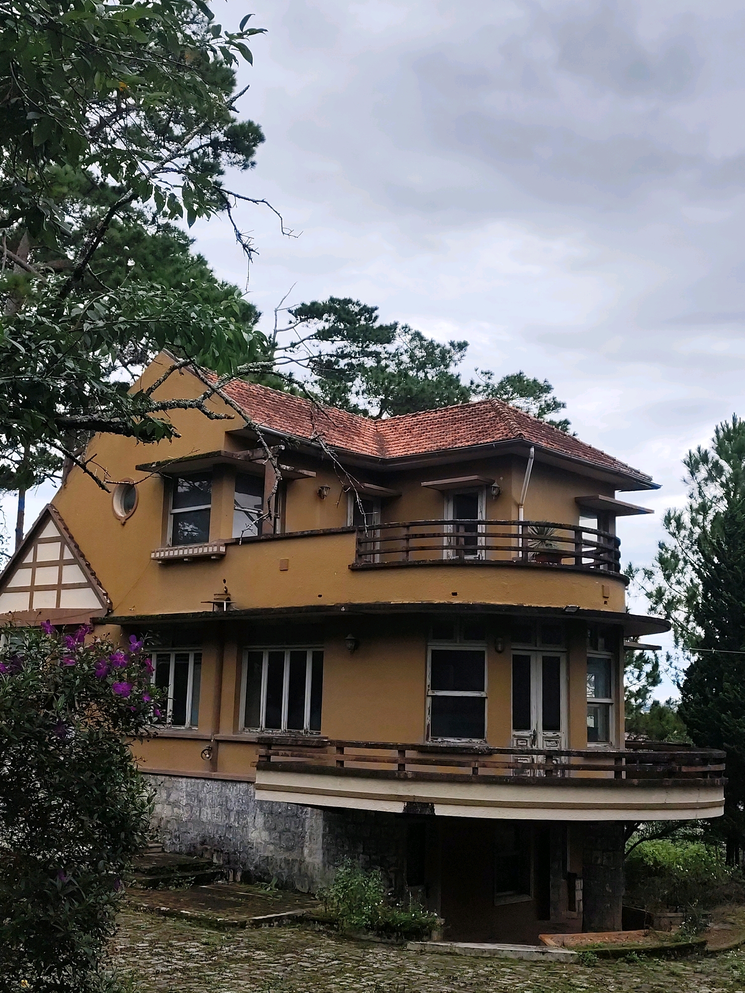 The image shows a two-story, mustard-yellow house with a red tile roof, surrounded by lush greenery and tall pine trees under a cloudy sky. The house features multiple balconies, large windows, and architectural details characteristic of French colonial style. In the foreground, a pathway made of cobblestones leads to the house, with a cluster of purple flowers on the left side. The house is situated in Da Lat, Vietnam, a city known for its French colonial architecture and temperate climate, often referred to as the "City of Eternal Spring." The scene evokes a sense of tranquility and rustic charm, hinting at the historical and natural beauty of the region.