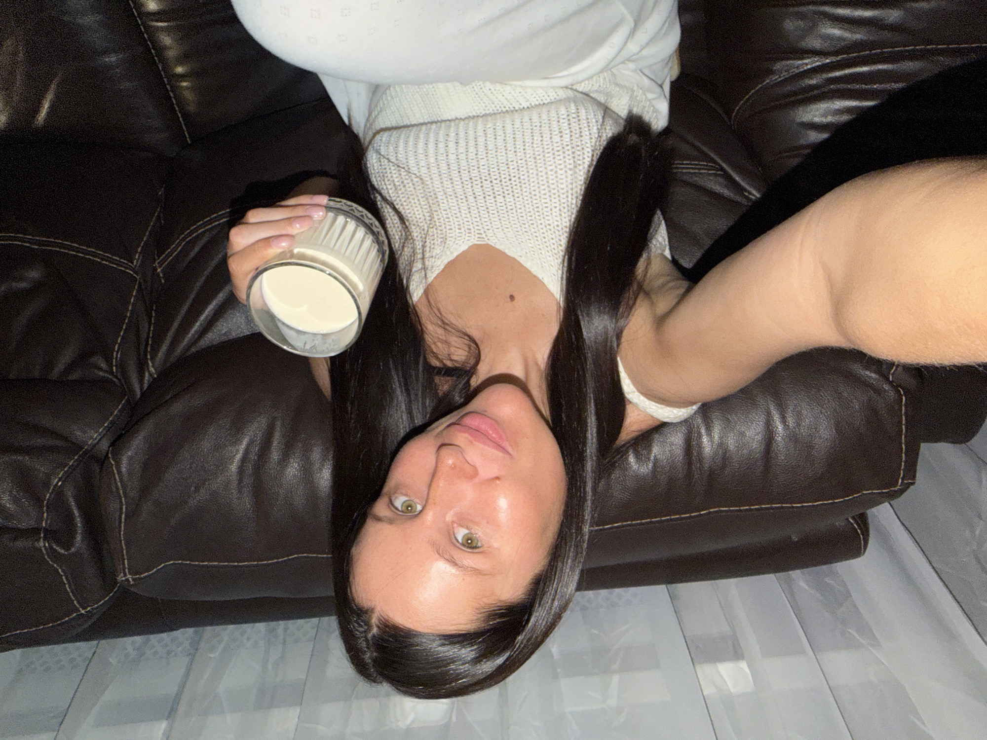 A woman is photographed lying upside down on a dark brown leather couch. She is holding a glass of white liquid in her left hand. The woman has long dark hair and is wearing a white top and light-colored pants. The background includes sheer white curtains. The location is unknown and cannot be determined as Khimki, Russia.