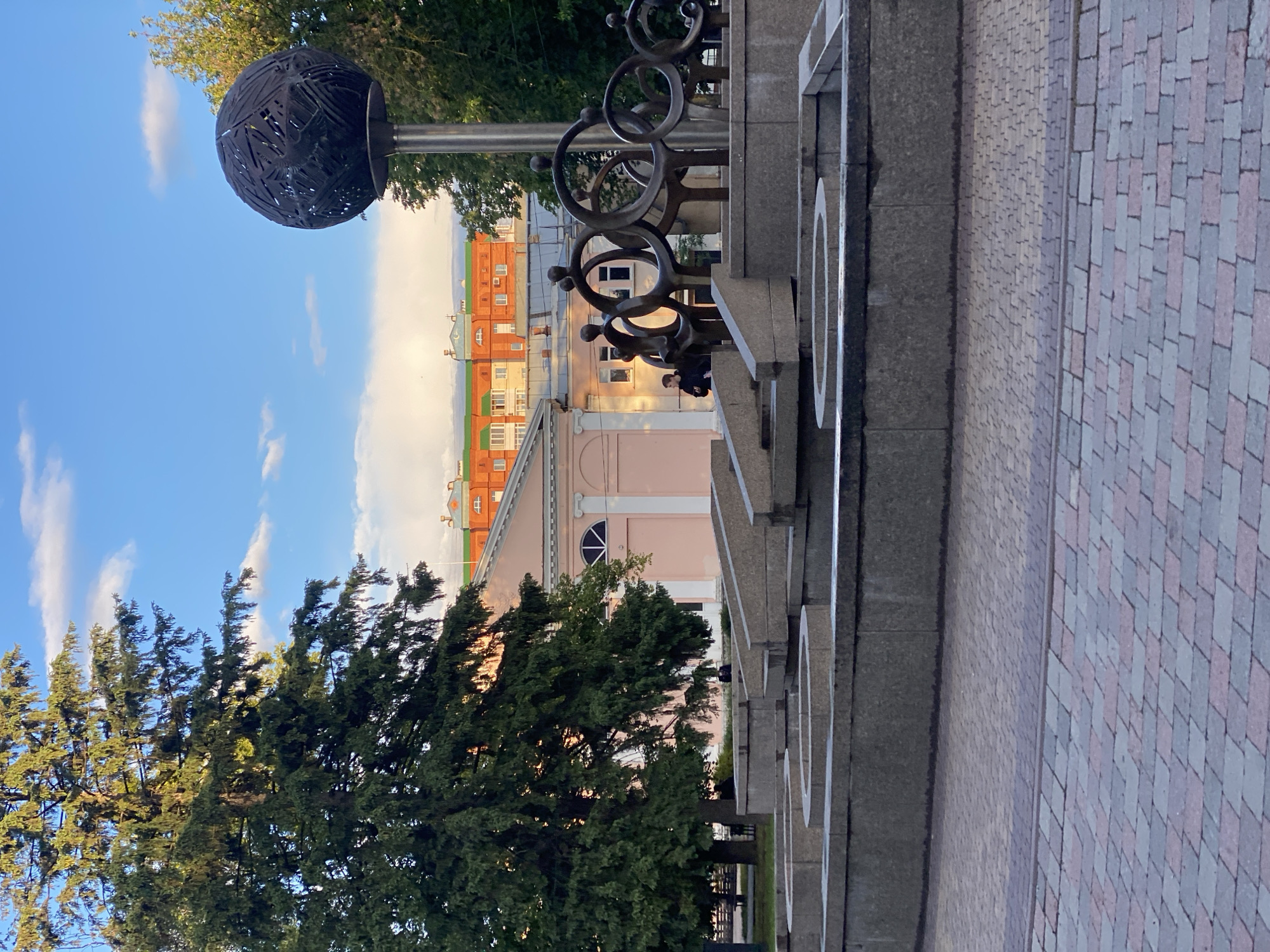 The image shows a section of a city park in Poltava, Ukraine. In the foreground, there is a stone and metal sculpture featuring interconnected rings and a person seated on the ledge of the sculpture. A decorative metal sphere atop a pole rises behind the sculpture. The park has trees on the left and a brick-paved walkway on the right. In the background are buildings, including one painted pink and another with orange and white bricks. The sky above has scattered clouds. The lighting indicates it is daytime.