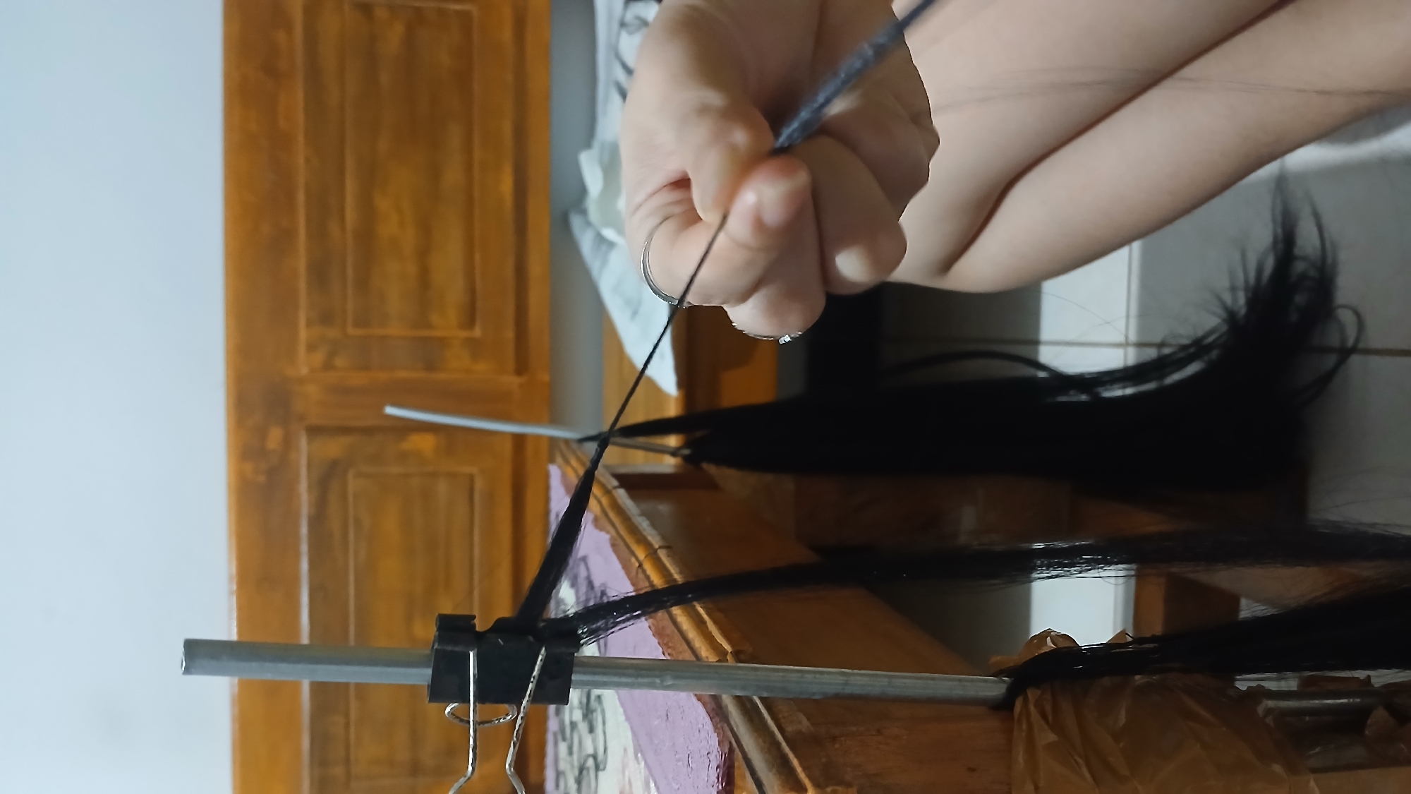 A close-up, waist-up shot shows a person's hands in the process of braiding dark hair. The hair is attached to a silver pole which is secured to a wooden surface with brown tape and a black clamp. The hands are positioned near the end of the hair, with one hand holding a thin, dark thread that is being woven into the braid. The background consists of a wooden headboard and a white wall. The scene is indoors, suggesting a home environment. The presence of the wooden furniture and the general lighting conditions are consistent with residential settings found in Sampang, Indonesia.
