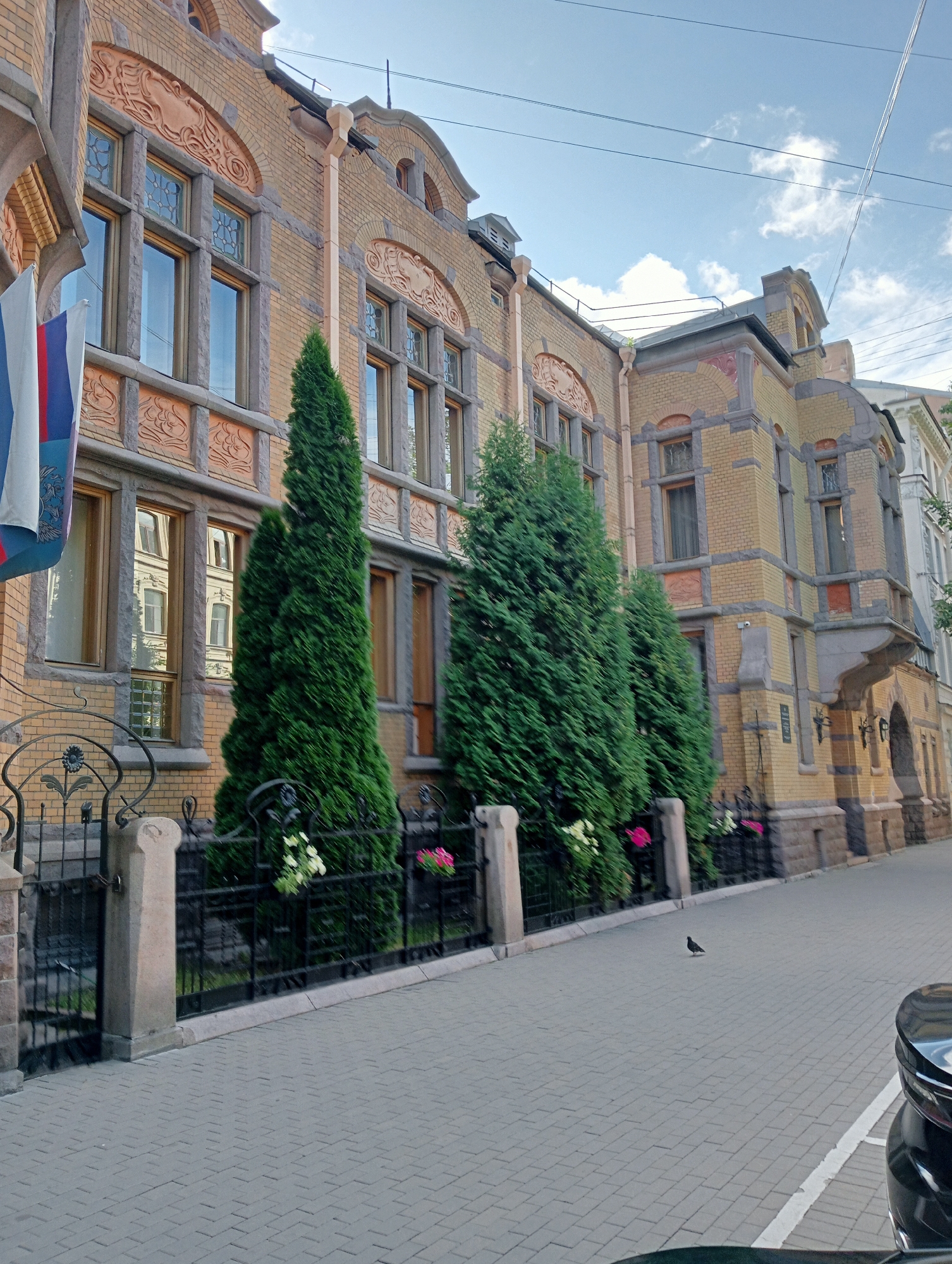 The image depicts a view of an ornate yellow brick building with a decorative façade, likely an embassy or a historic institution, situated on a street in Saint Petersburg, Russia. The building features numerous tall windows with stained glass and intricate stone carvings above them. A black metal fence with decorative elements separates the building's grounds from a paved sidewalk. Several tall, dark green evergreen trees are planted behind the fence. Two flags, one white and blue with a red stripe, and another predominantly white with blue and red accents, are visible on the left side of the building. A small black bird is perched on the sidewalk in the middle ground. The sky is light blue with scattered white clouds, and power lines are visible in the upper portion of the frame. A portion of a black car is visible in the lower right corner. The lighting suggests it is daytime.