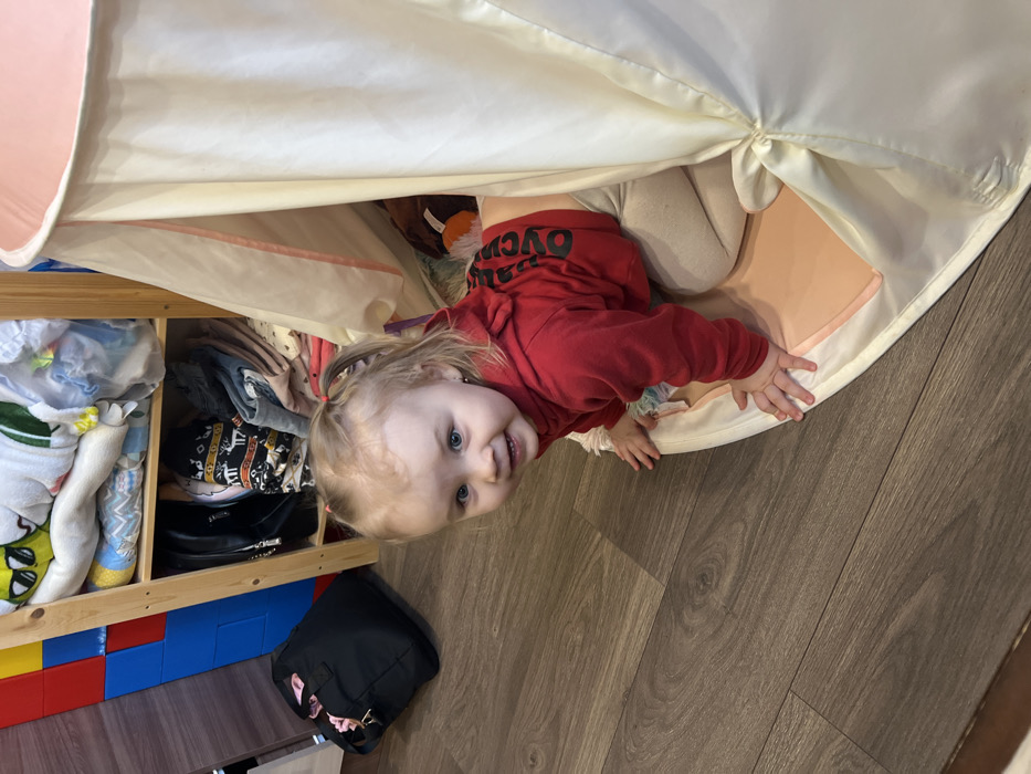 A young child with blonde hair tied in two pigtails and distinct blue eyes is depicted smiling and looking directly towards the viewer. The child wears a red long-sleeved hooded shirt with black Cyrillic text visible on the back, and light-colored leggings, positioned on hands and knees. The child is partially emerging from the opening of a cream-colored fabric teepee, which has a circular pink base pad. In the background, a light-toned wooden shelving unit contains folded textiles, including blankets with patterns, on its top shelf, and a black handbag alongside other garments on its lower shelf. To the left of the shelving unit, a black fabric tote bag rests on dark brown laminate wood flooring. Adjacent to the shelving unit and behind the bag, a wall section is covered with large, colorful modular panels in red, blue, and yellow. This indoor scene is located in Leninakan, Russia.