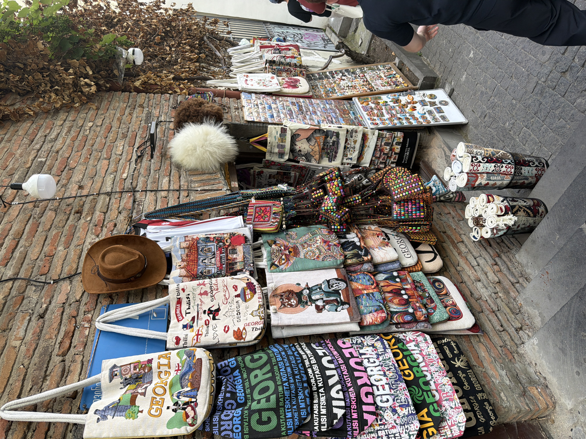 A street vendor stall in Tbilisi, Georgia, displays a variety of souvenirs. The stall is situated against a brick wall and on a stone staircase. Products include embroidered tote bags with "GEORGIA" and images of local landmarks, a brown cowboy hat, fluffy hats, small decorative items, and rolled-up textiles in cylindrical displays. A person in a black t-shirt and pants is visible on the right side of the frame, walking away from the stall. The environment appears to be an outdoor market or shopping street in a historic district.